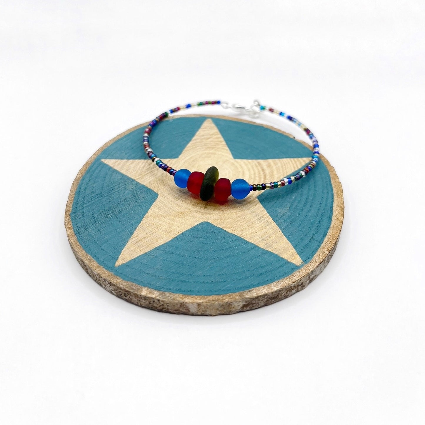 A rainbow coloured beaded bracelet with red and blue frosted glass beads and a piece of olive green coloured Cornish sea glass, finished with a silver plated lobster clasp and laid on a slice of wood with a blue star design.