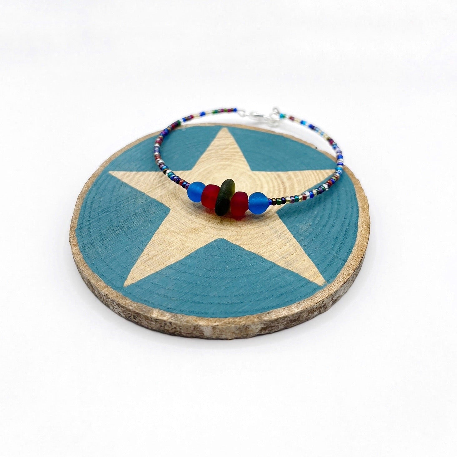 A rainbow coloured beaded bracelet with red and blue frosted glass beads and a piece of olive green coloured Cornish sea glass, finished with a silver plated lobster clasp and laid on a slice of wood with a blue star design.