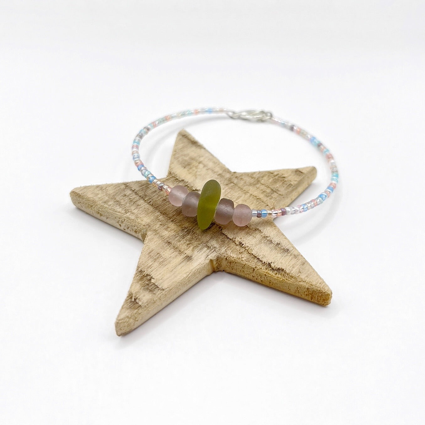 A pastel beaded bracelet with purple and lilac frosted glass beads and a piece of soft lime coloured Cornish sea glass, finished with a silver plated lobster clasp and laid on a wooden star.