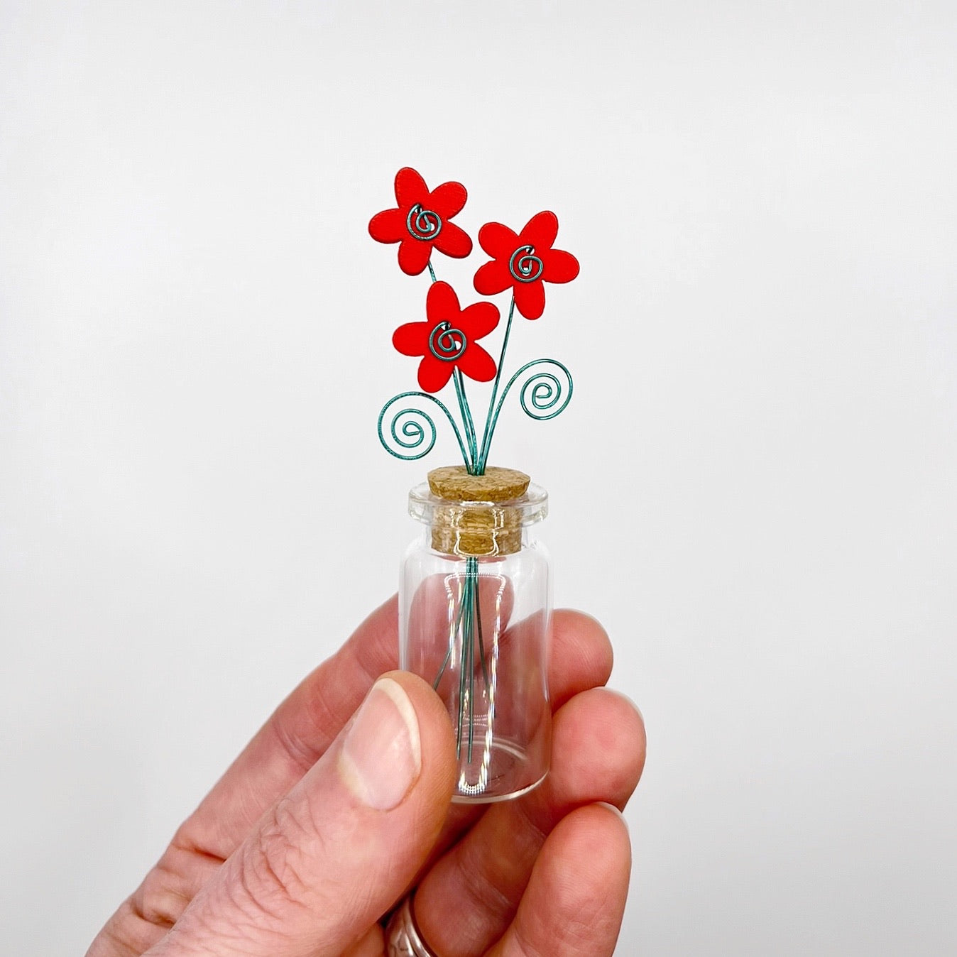 Hand holding a small glass bottle with red wooden flowers against a white background
