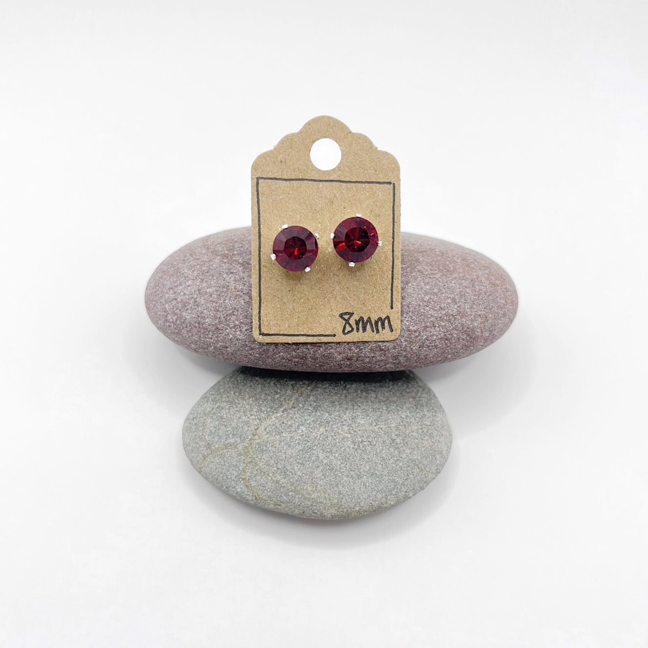 Pair of red cut glass stud earrings on a cardboard backing with '8mm' text, placed on two stones.