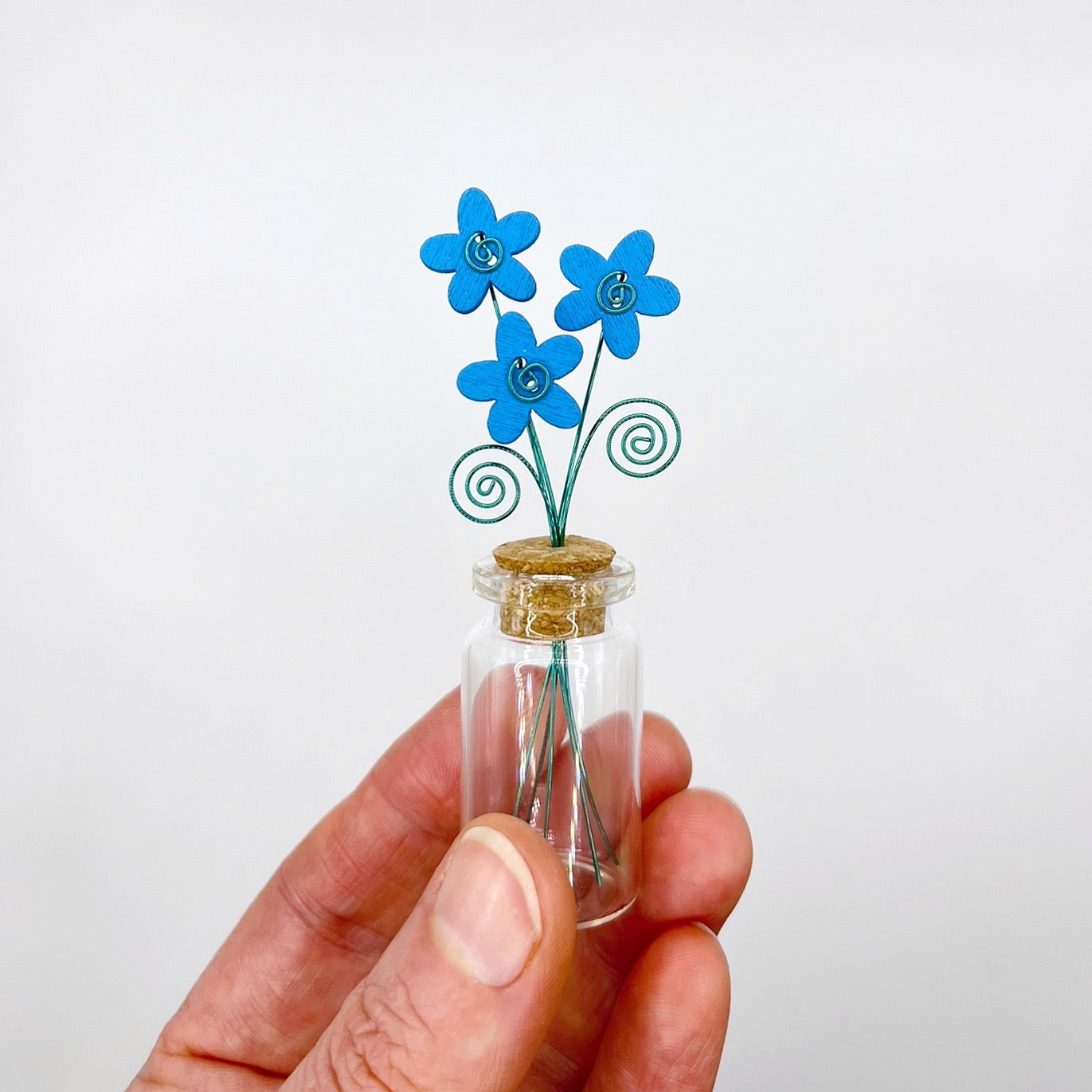 Hand holding a small glass bottle with blue wooden flowers against a white background