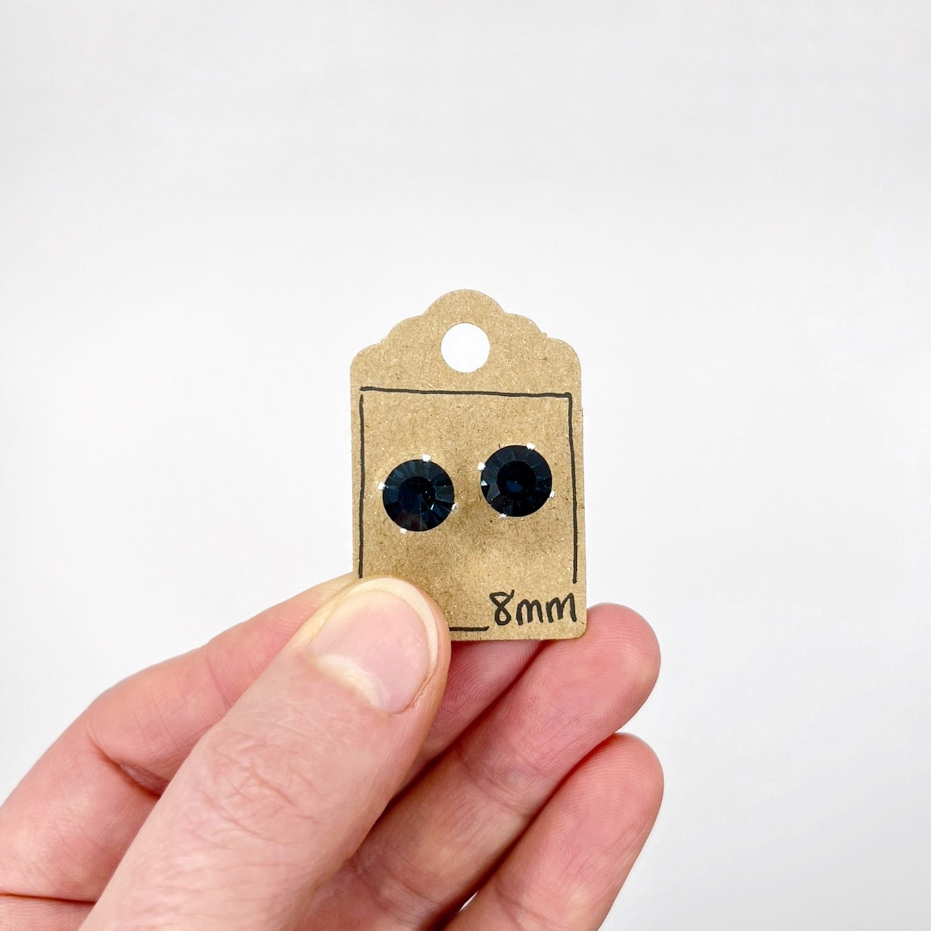 Blue cut glass stud earrings on a cardboard tag held by a hand against a white background