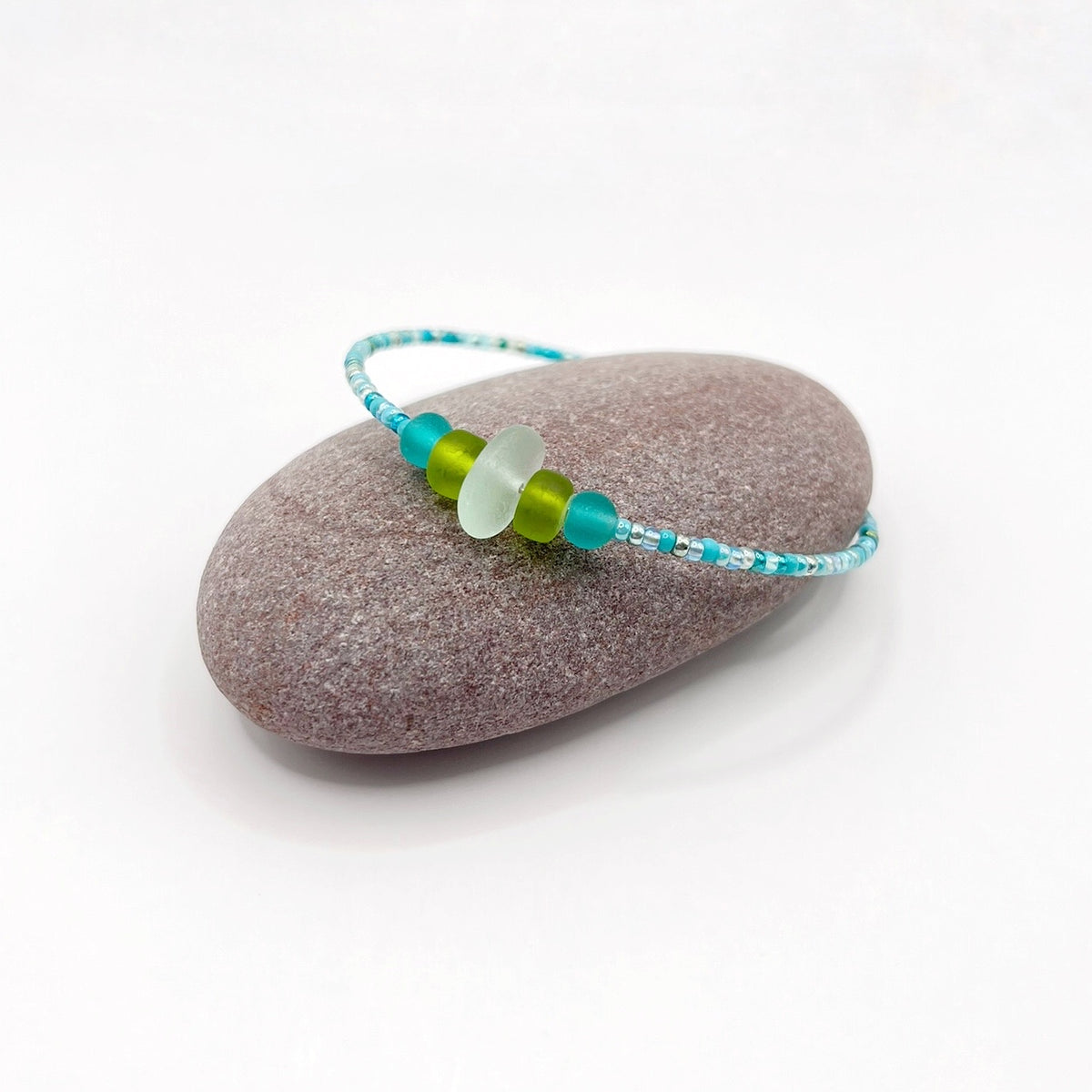 A turquoise beaded bracelet with lime and sea green frosted glass beads and a piece of sea foam coloured Cornish sea glass, finished with a silver plated lobster clasp and laid on a purple pebble.