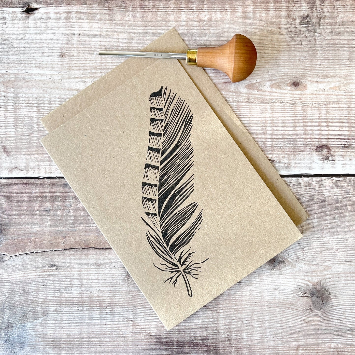 Feather lino print in black on a kraft card with a matching envelope. Photo includes lino cutting tool used.