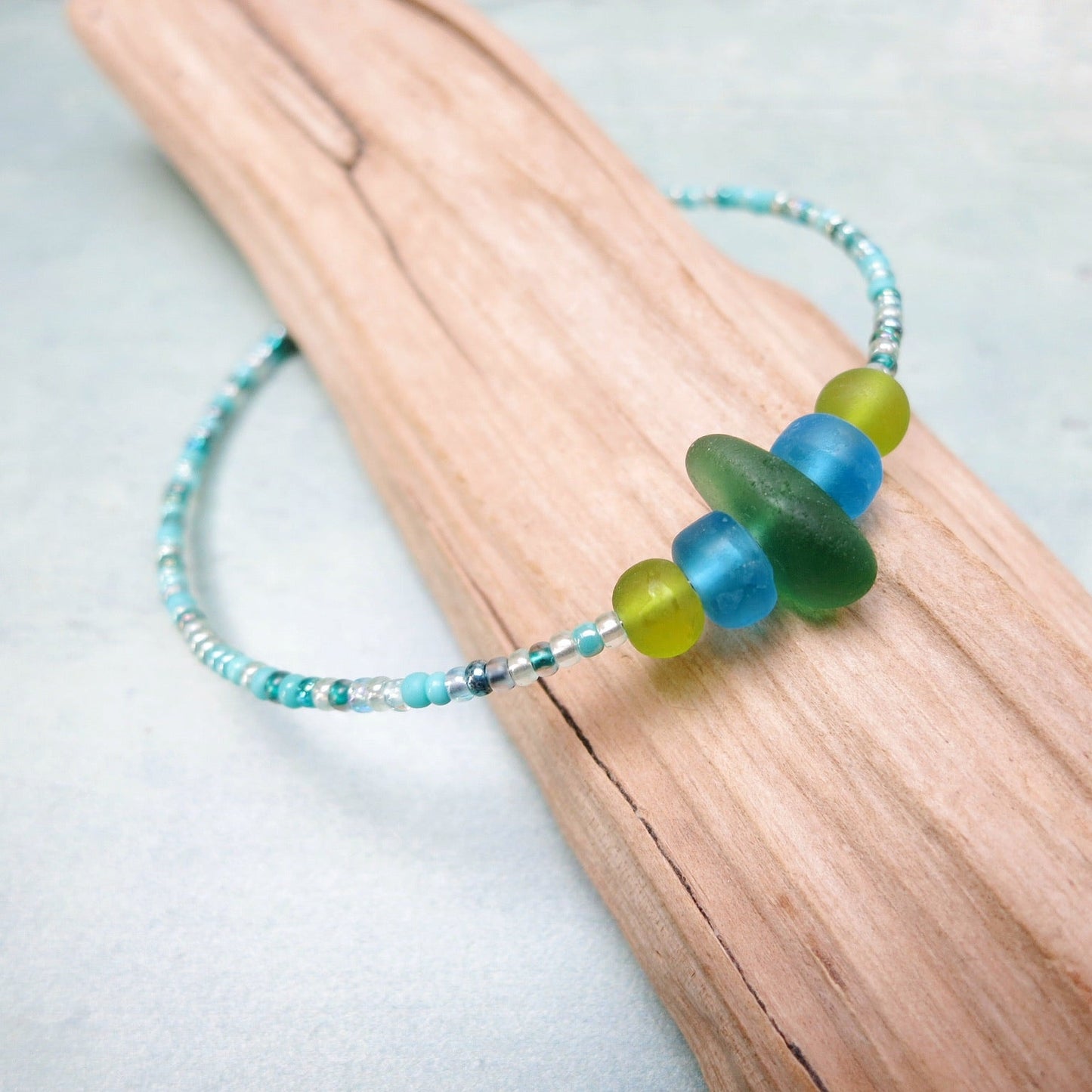 Bracelet with green sea glass and turquoise and green glass beads laid on driftwood against a pale blue background