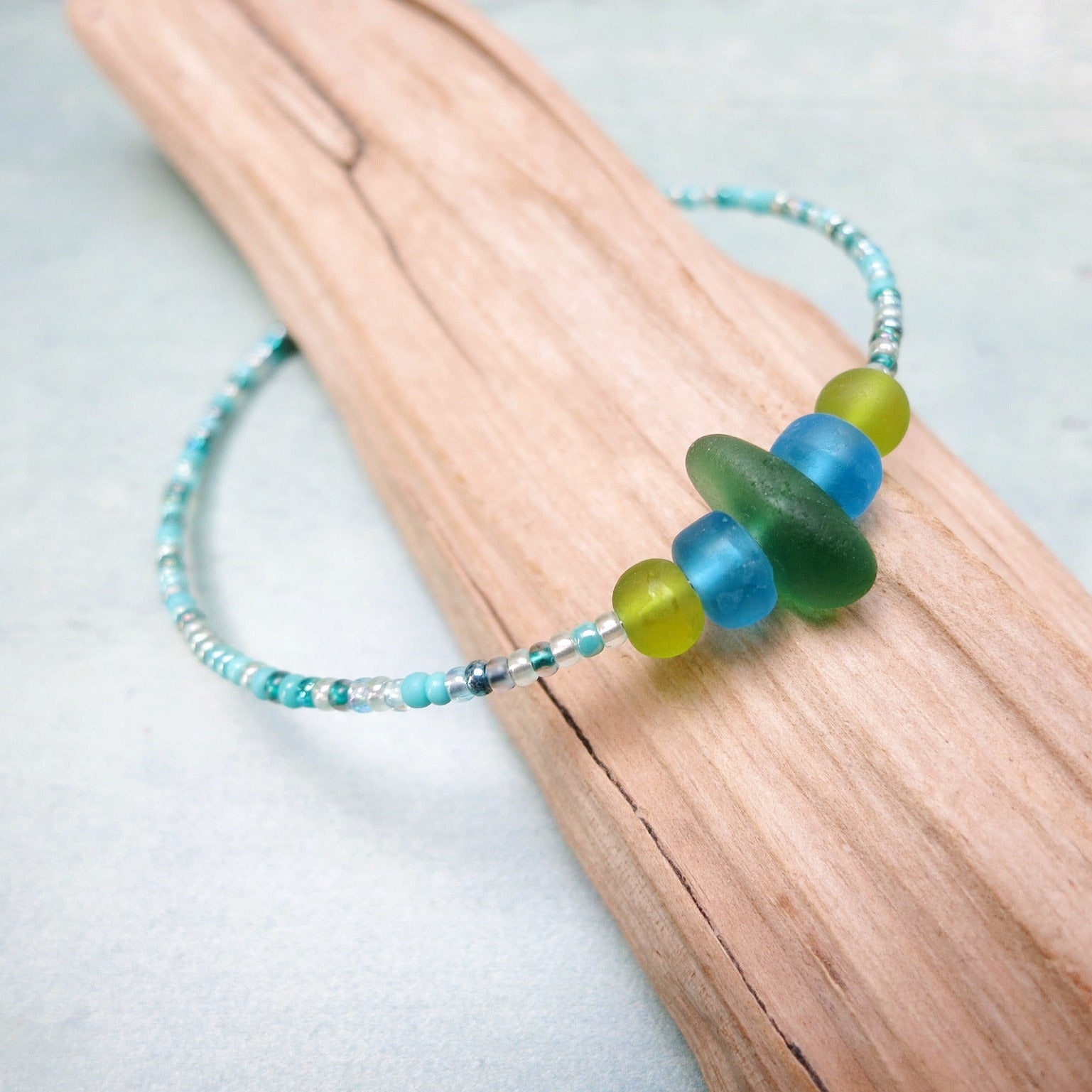 Bracelet with green sea glass and turquoise and green glass beads laid on driftwood against a pale blue background