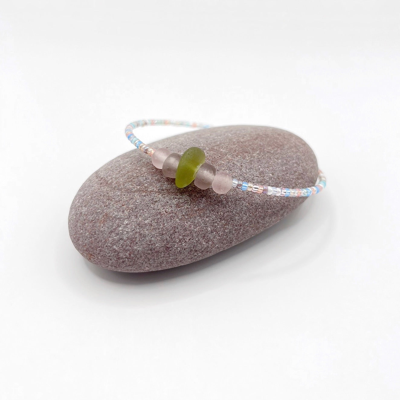 A pastel beaded bracelet with purple and lilac frosted glass beads and a piece of soft lime coloured Cornish sea glass, finished with a silver plated lobster clasp and laid on a purple pebble.