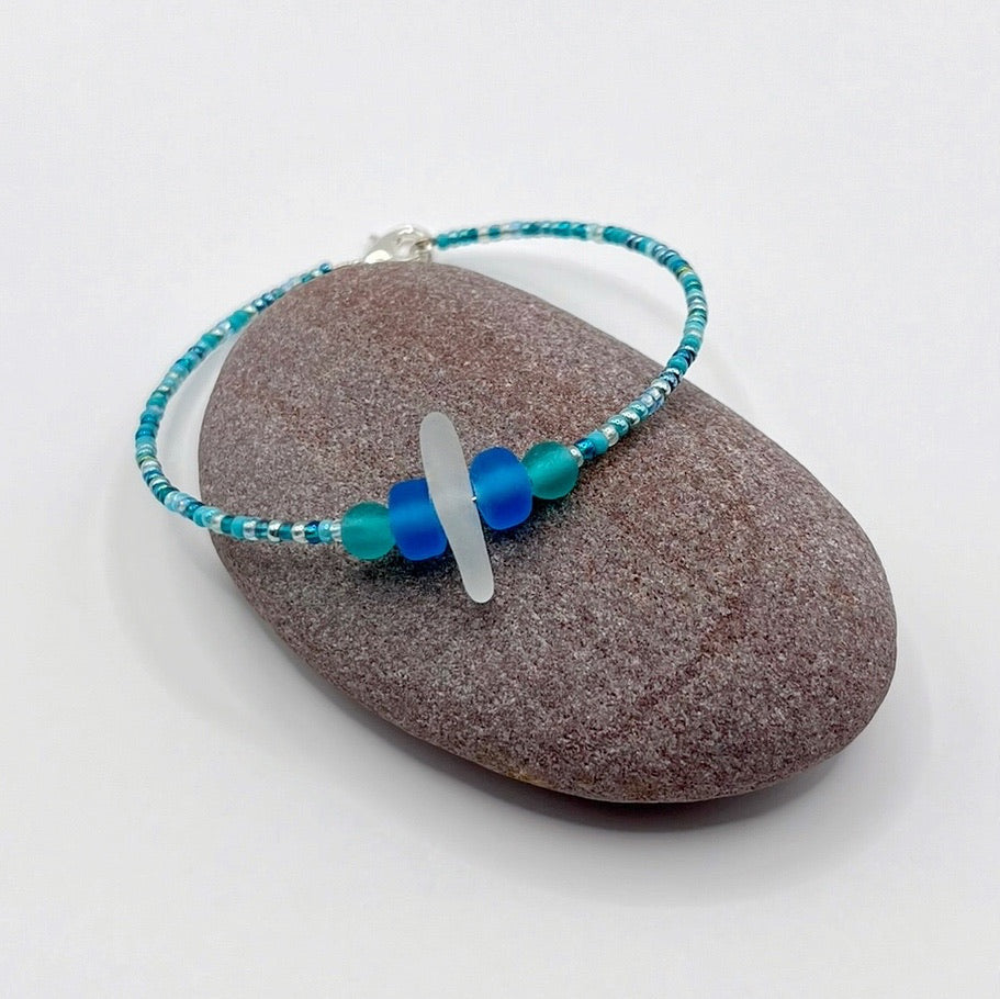Bracelet with white sea glass and blue and turquoise beads laid on a purple pebble against a white background