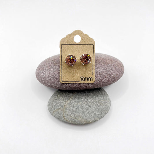Pair of cut glass stud earrings on a cardboard backing with '8mm' marking, placed on two stones.