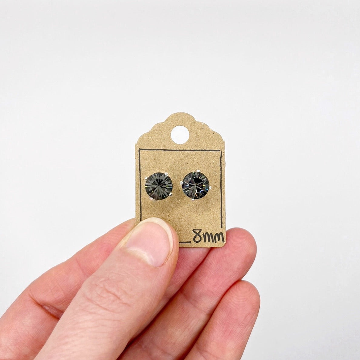 Hand holding a small cardboard earring display card with grey cut glass stud earrings on a white background