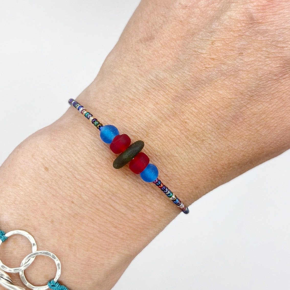 Close up of a rainbow coloured beaded bracelet with red and blue frosted glass beads and a piece of olive green coloured Cornish sea glass, worn on a wrist.