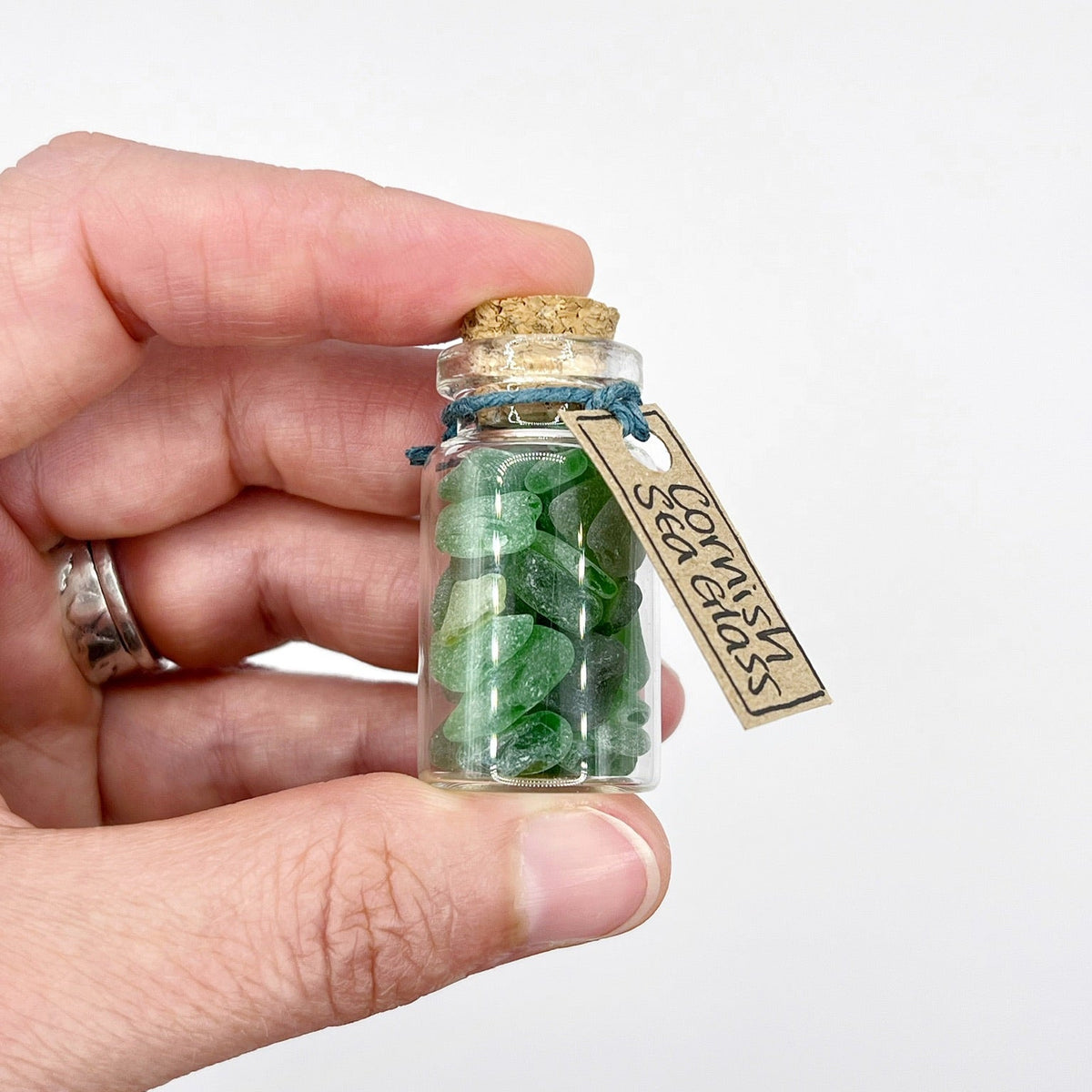 A hand holding a small glass bottle filled with green sea glass, sealed with a cork and tied with blue string, featuring a hanging tag that reads 'Cornish Sea Glass'.