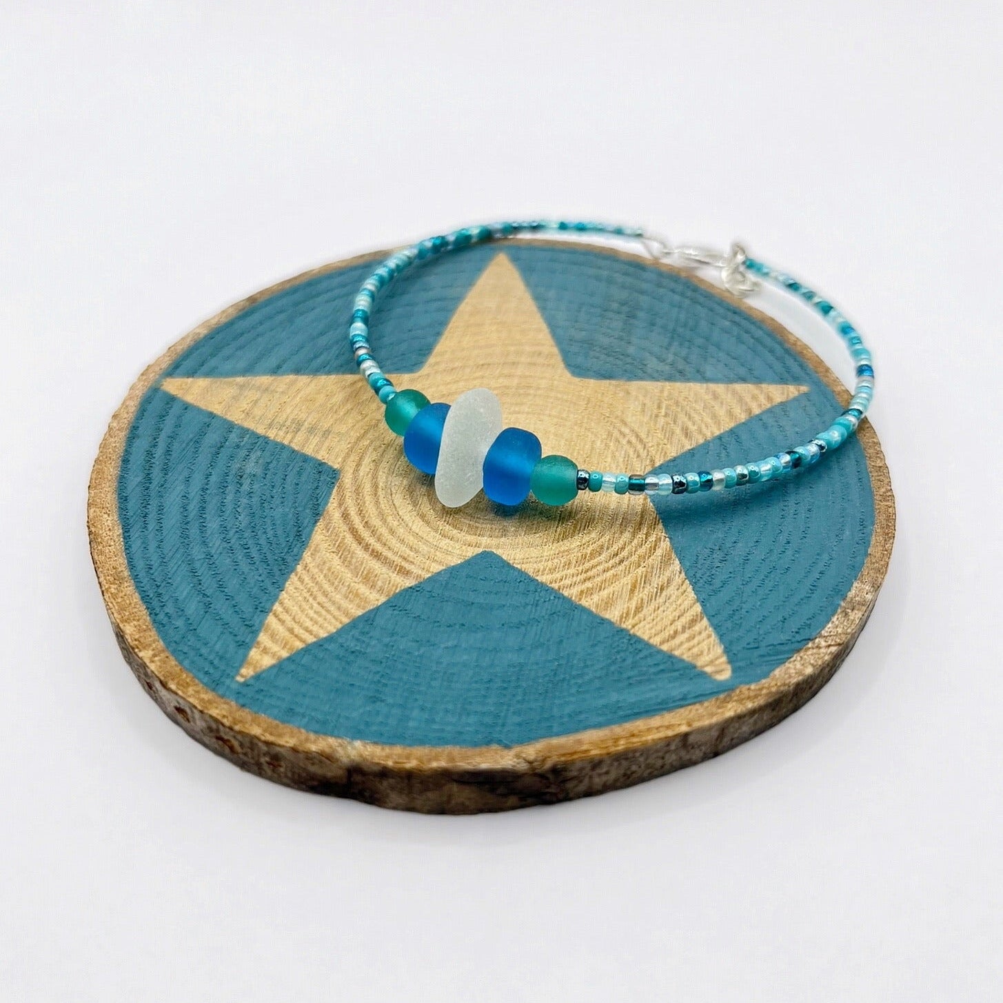 Bracelet with white sea glass and blue and turquoise beads laid on a slice of wood with a blue painted star shape against a white background