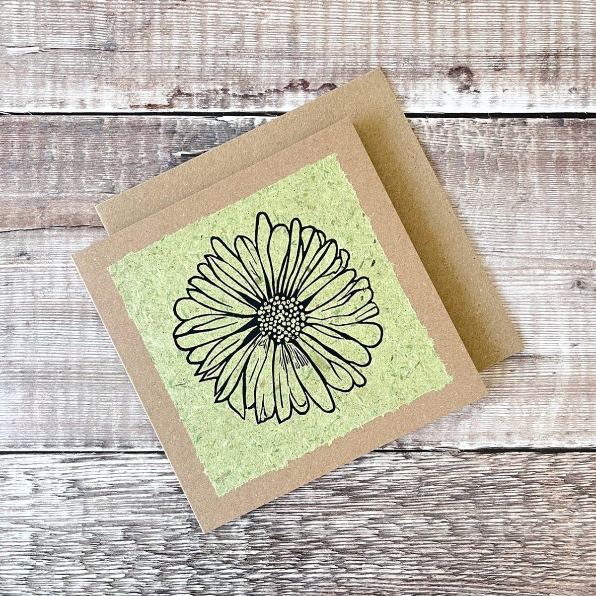 A square handmade art card featuring a black ink lino print of a daisy flower on a textured light green handmade paper, mounted on a brown kraft greetings card, laid flat on a rustic wooden surface.