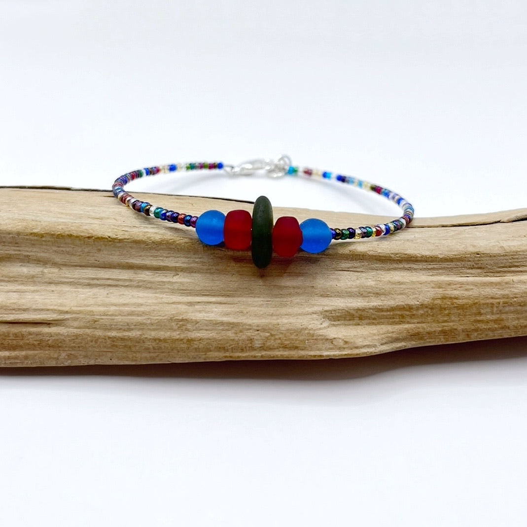 A rainbow coloured beaded bracelet with red and blue frosted glass beads and a piece of olive green coloured Cornish sea glass, finished with a silver plated lobster clasp and laid on a piece of driftwood.