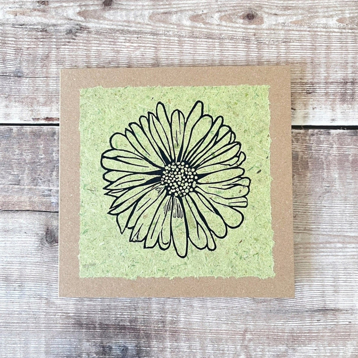 A square handmade art card featuring a black ink lino print of a daisy flower on a textured light green handmade paper, mounted on a brown kraft greetings card, laid flat on a rustic wooden surface.