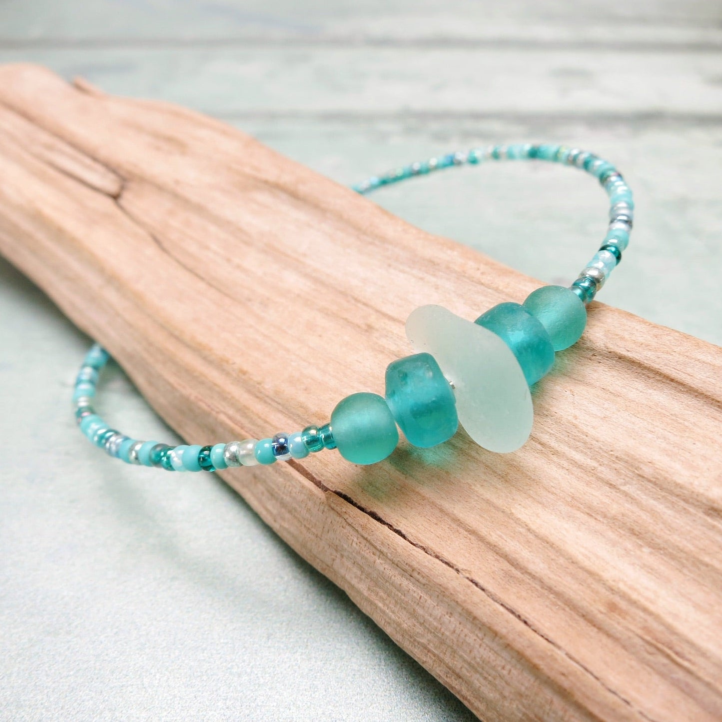 Bracelet with pale sea foam sea glass and sea green and turquoise glass beads laid on driftwood against a pale blue background