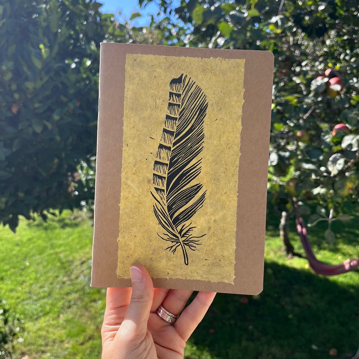 A hand holding an A5 kraft paper notebook or journal with a textured yellow handmade paper panel featuring a black lino ink feather illustration, photographed against a garden backdrop.