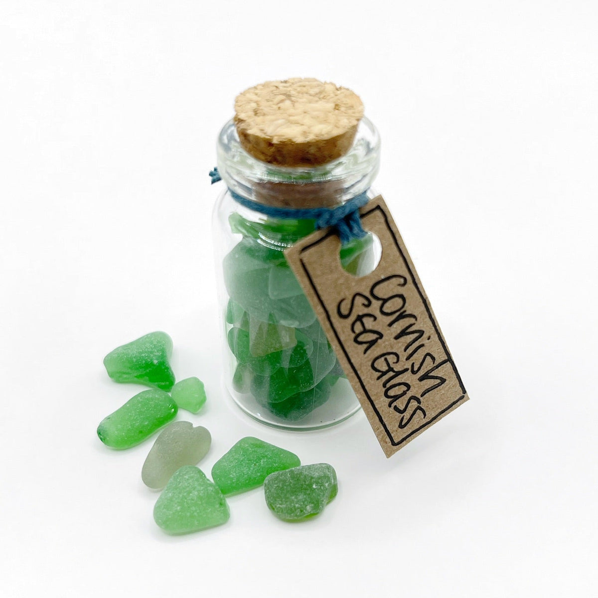 A small clear glass bottle with a cork stopper and blue string, filled with green sea glass, with several loose green sea glass pieces in front. A paper tag reads 'Cornish Sea Glass'.