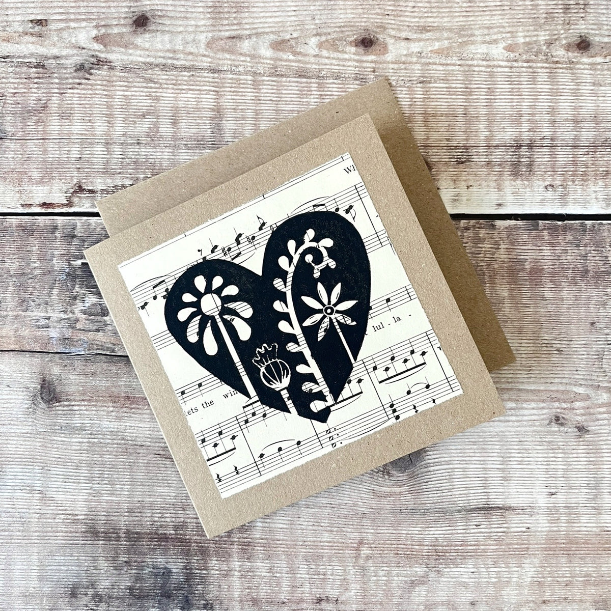 Square kraft greeting card with a black floral heart lino print on vintage music paper mounted on the front. Laid on a matching envelope on a wooden background.