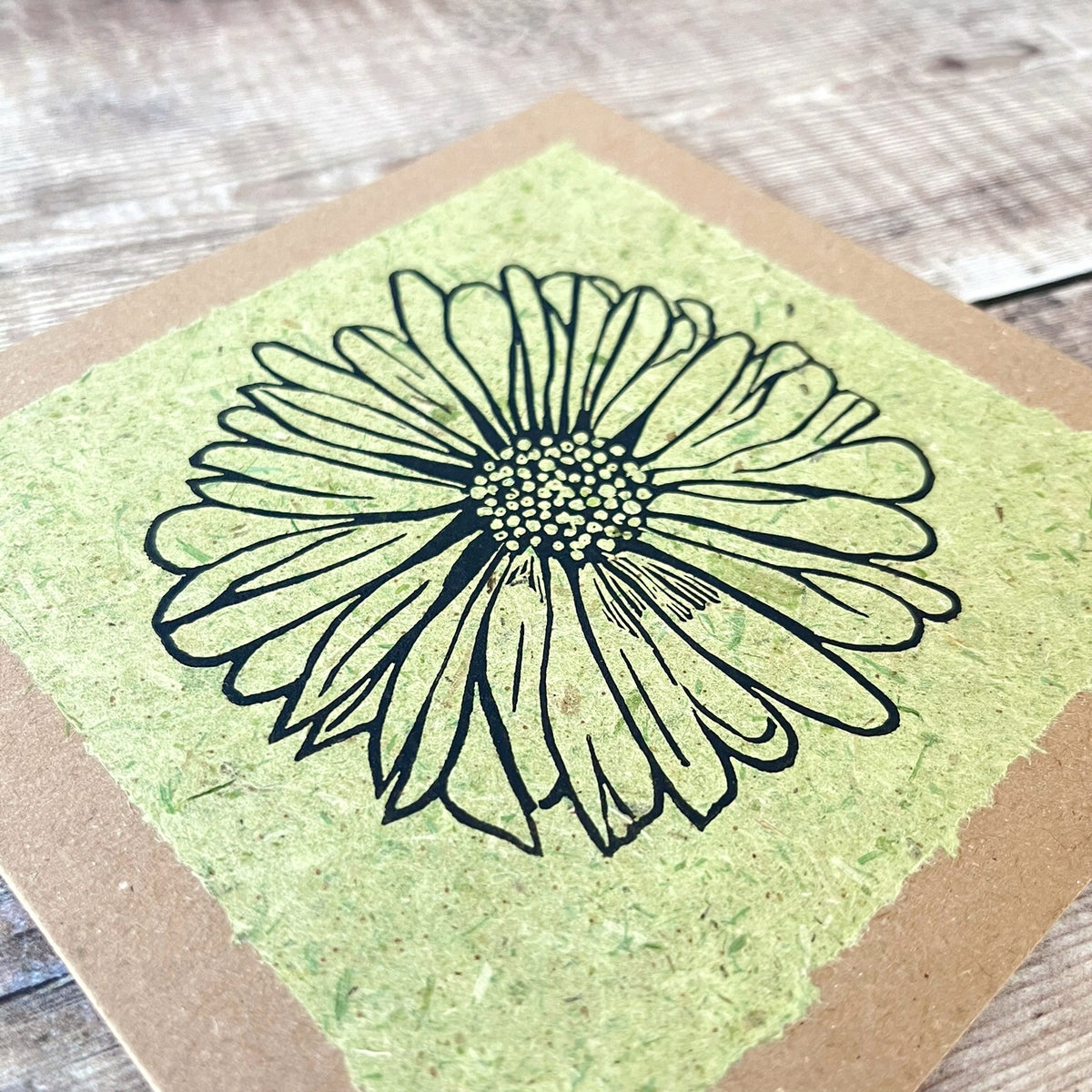Close up of a square handmade art card featuring a black ink lino print of a daisy flower on a textured light green handmade paper, mounted on a brown kraft greetings card, laid flat on a rustic wooden surface.