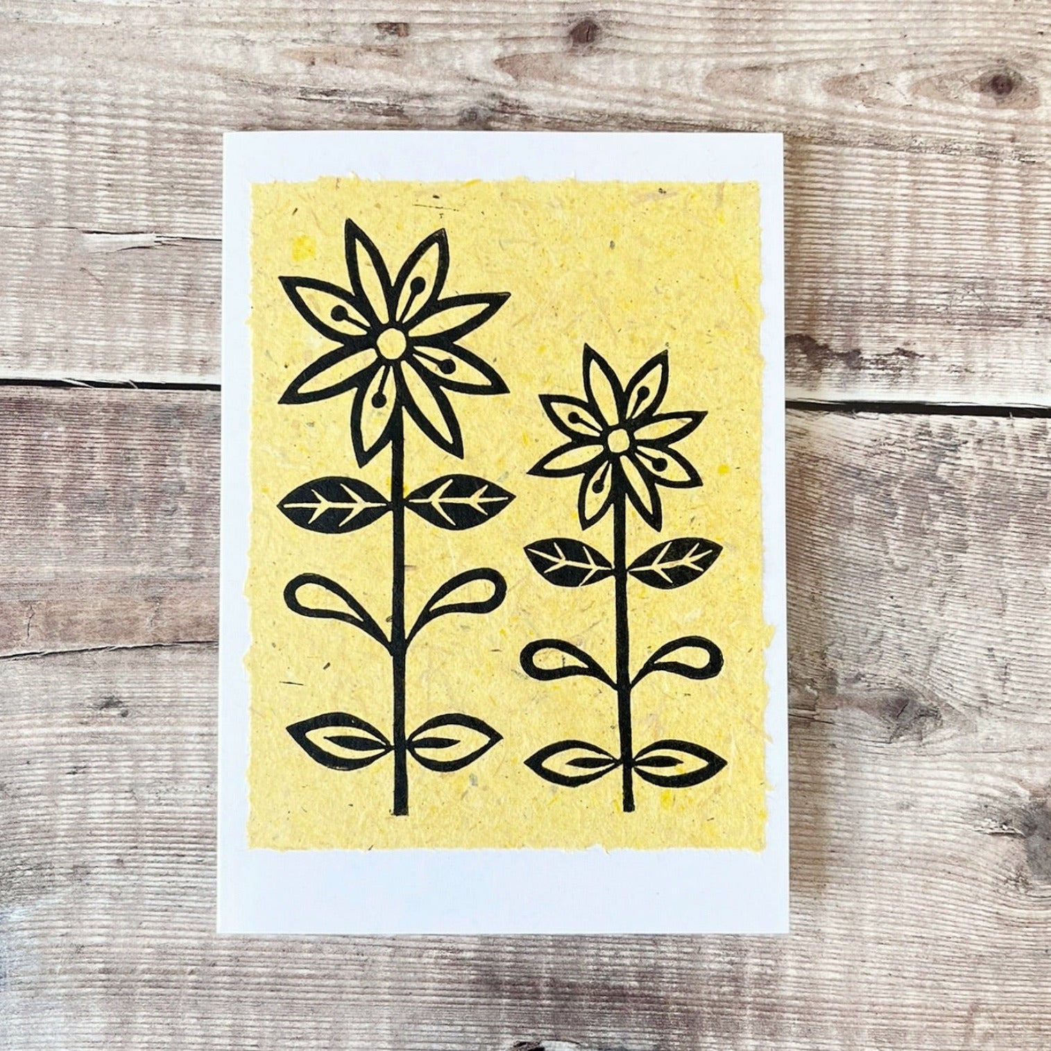 Lino print of two daisies in black ink on yellow handmade paper mounted on a white greetings card. Laid on a wooden surface.