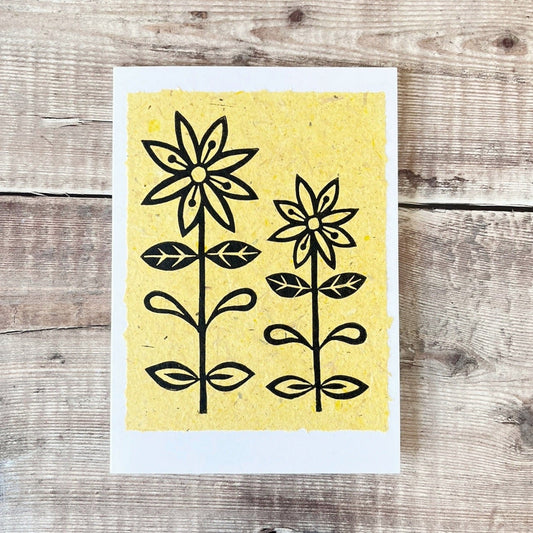 Lino print of two daisies in black ink on yellow handmade paper mounted on a white greetings card. Laid on a wooden surface.