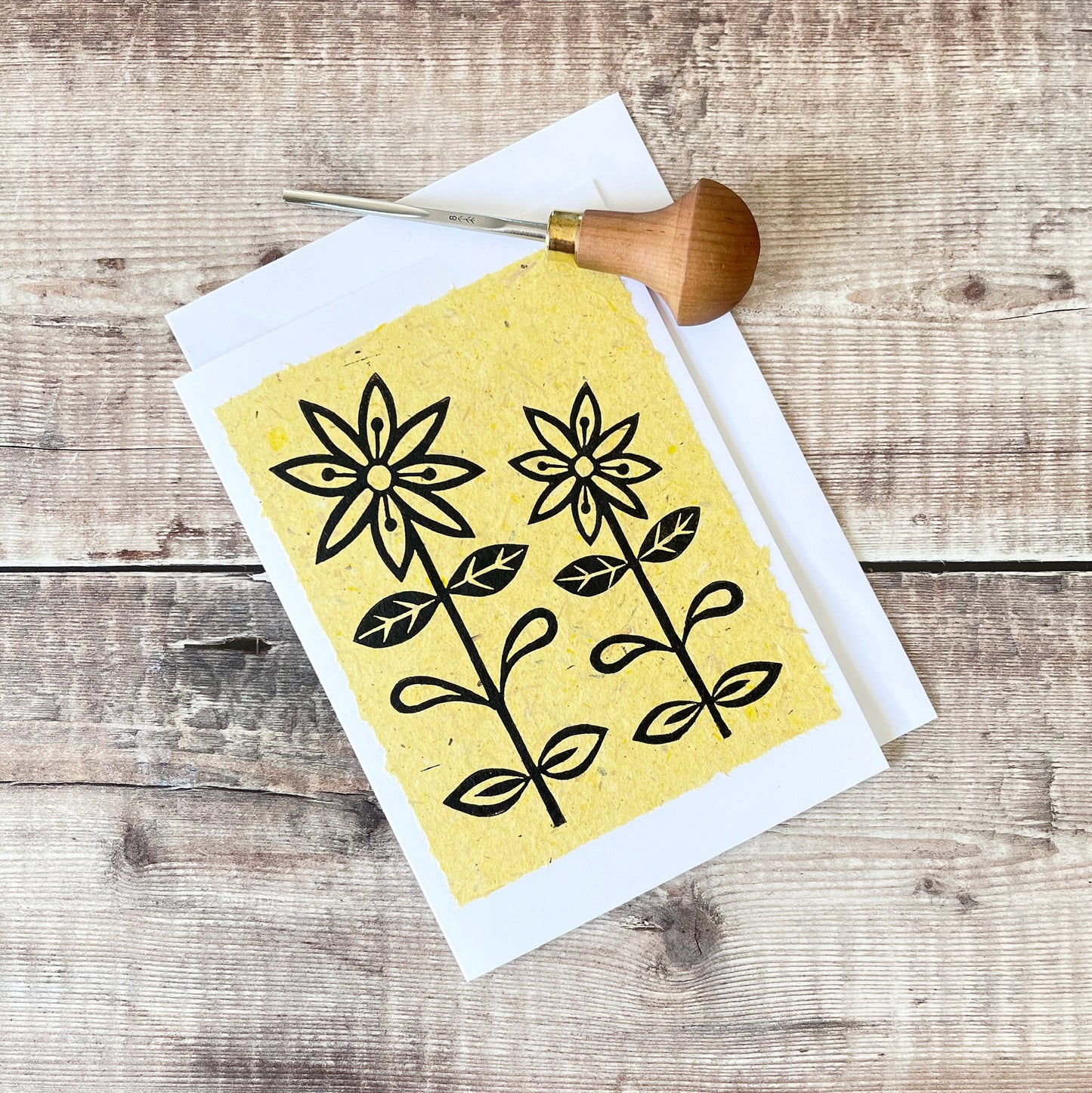 Lino print of two daisies in black ink on yellow handmade paper mounted on a white greetings card with matching envelope. Photo includes cutting tool and is laid on a wooden surface.