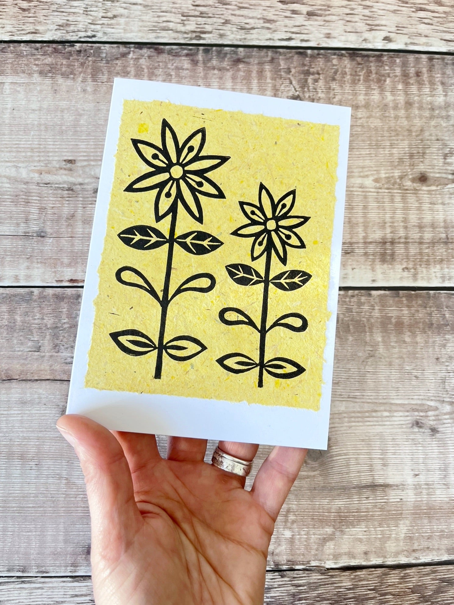 Lino print of two daisies in black ink on yellow handmade paper mounted on a white greetings card. Held in a hand against a wooden surface.