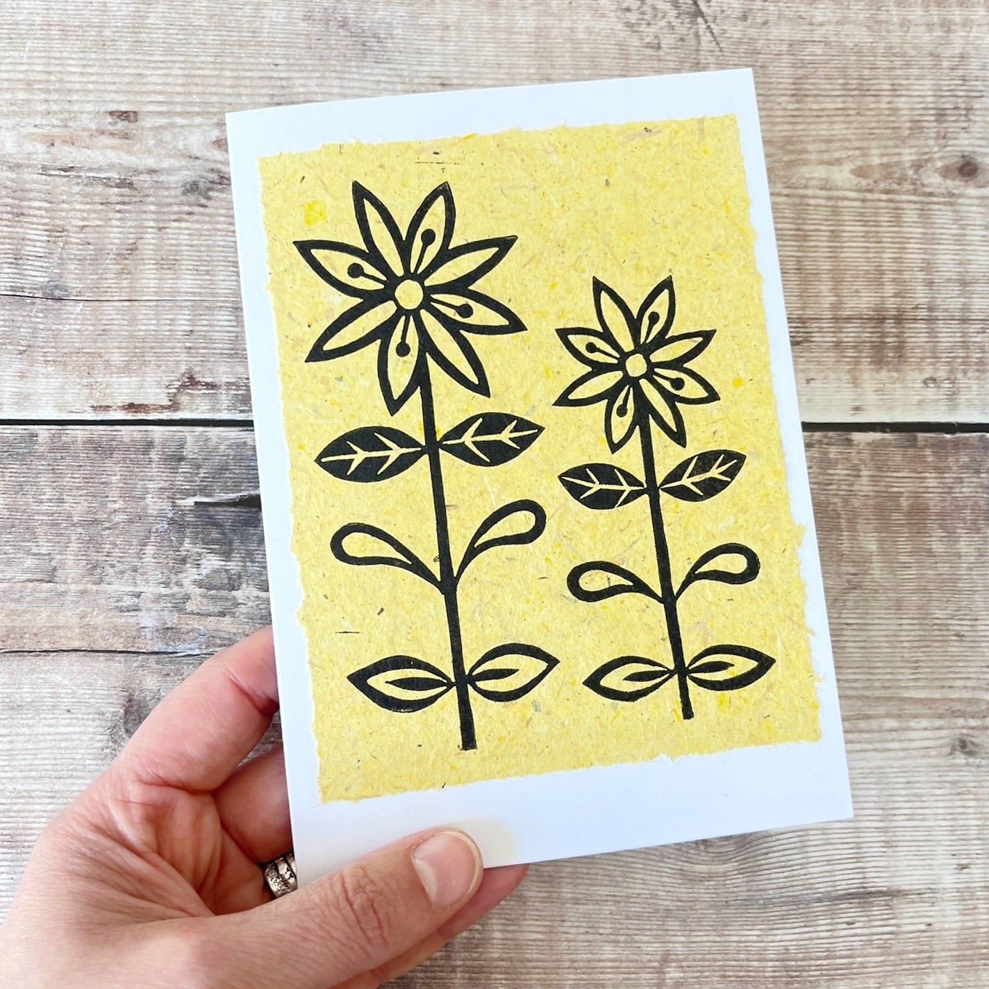 Lino print of two daisies in black ink on yellow handmade paper mounted on a white greetings card. Held in a hand against a wooden surface.