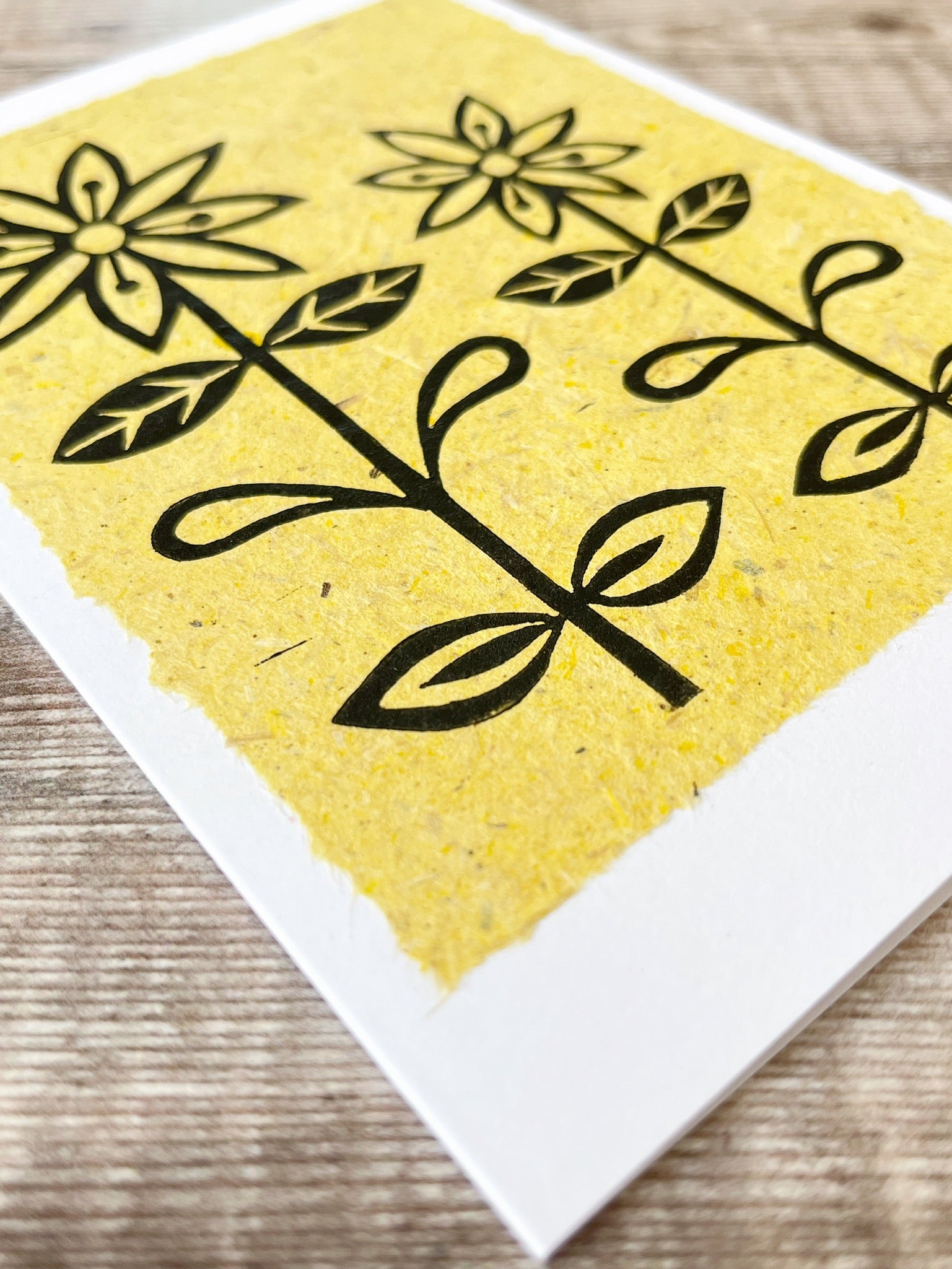 Close up of lino print of two daisies in black ink on yellow handmade paper mounted on a white greetings card. Laid on a wooden surface.