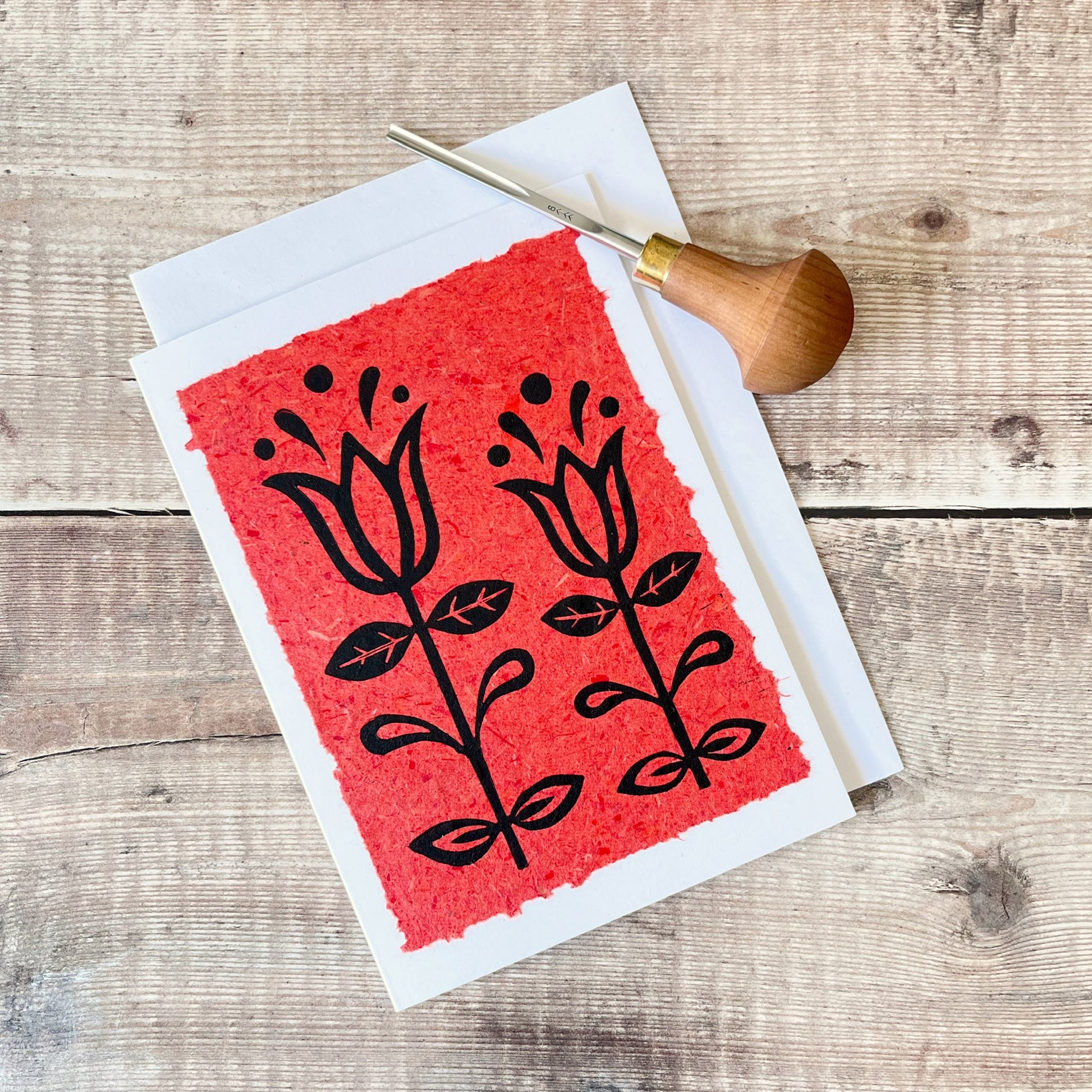 Lino print of two tulips in black ink on brick red handmade paper mounted on a white greetings card with matching envelope. Photo includes cutting tool and is laid on a wooden surface.