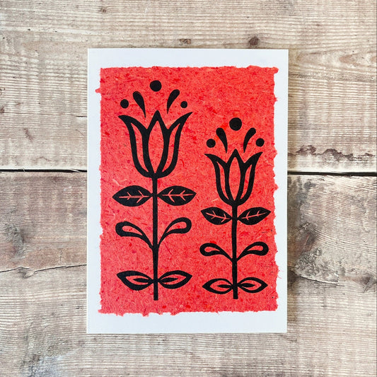 Lino print of two tulips in black ink on brick red handmade paper mounted on a white greetings card. Laid on a wooden surface.