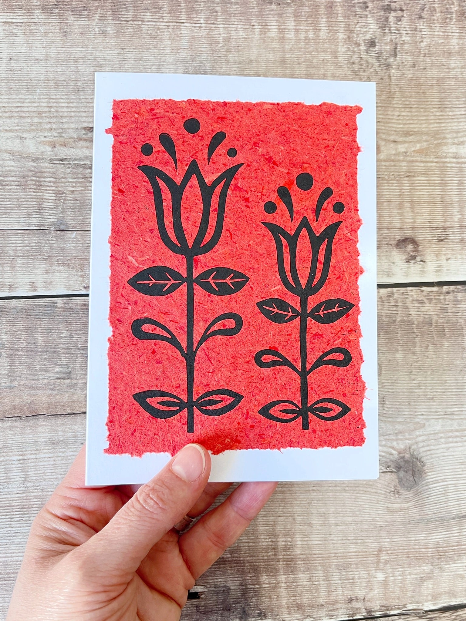 Lino print of two tulips in black ink on brick red handmade paper mounted on a white greetings card. Held in a hand against a wooden surface.