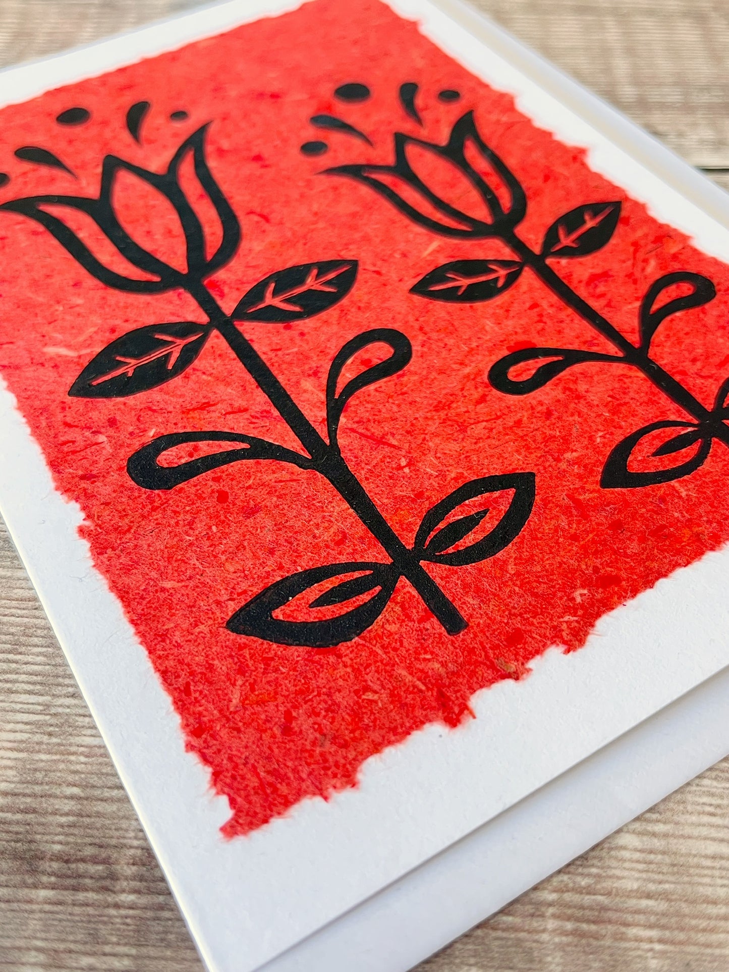 Close up of lino print of two tulips in black ink on brick red handmade paper mounted on a white greetings card. Laid on a wooden surface.