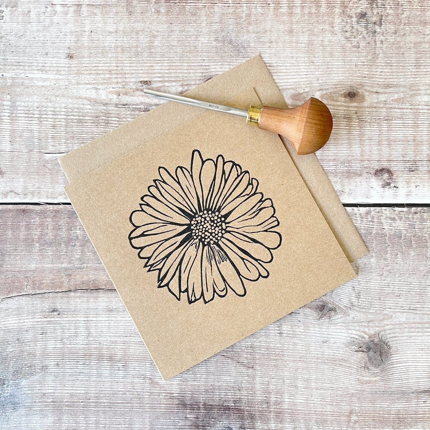Black lino printed Daisy design on kraft card with a matching envelope laid on a wooden surface. Photo includes lino cutting tool used.