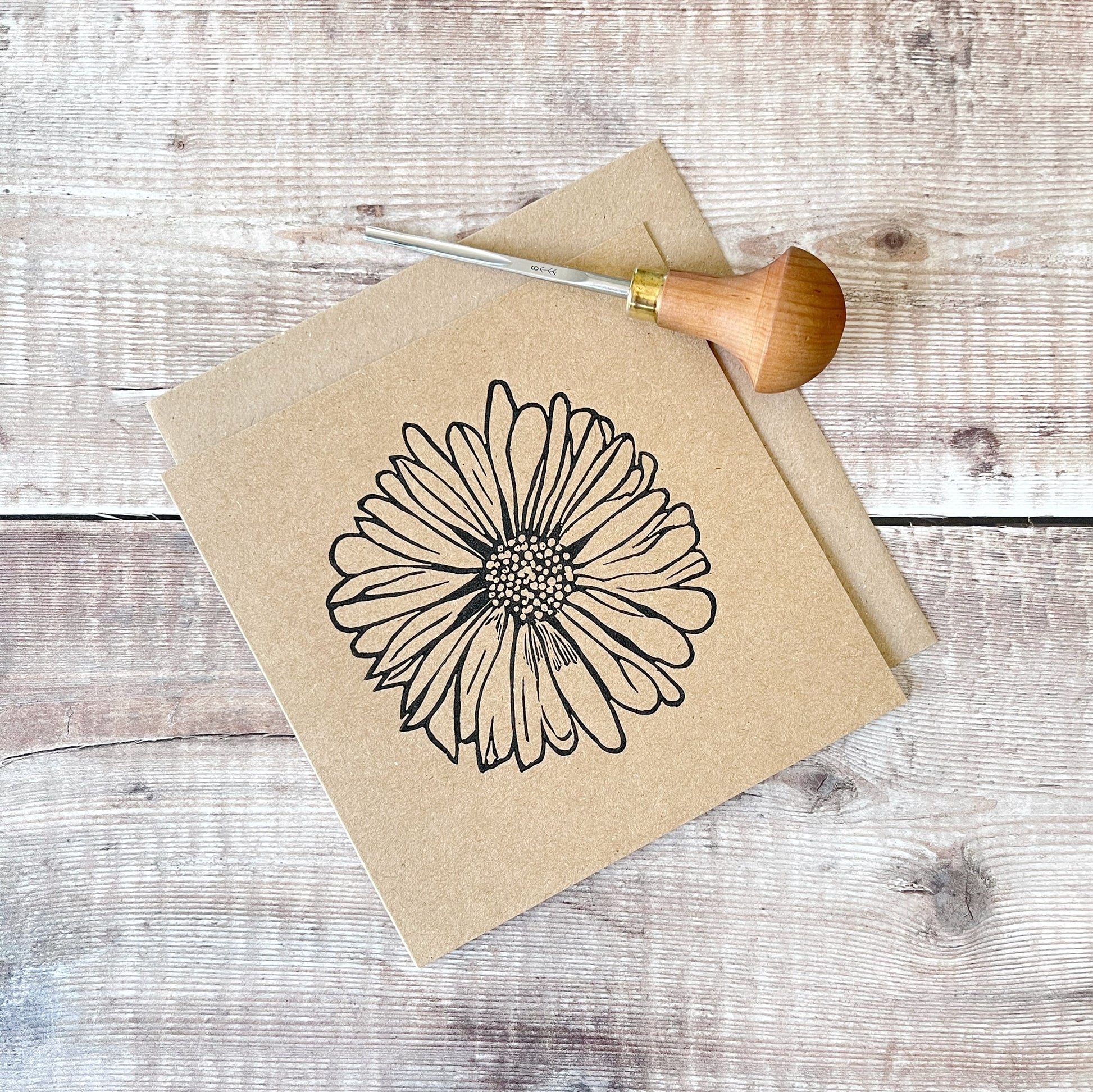 Black lino printed Daisy design on kraft card with a matching envelope laid on a wooden surface. Photo includes lino cutting tool used.