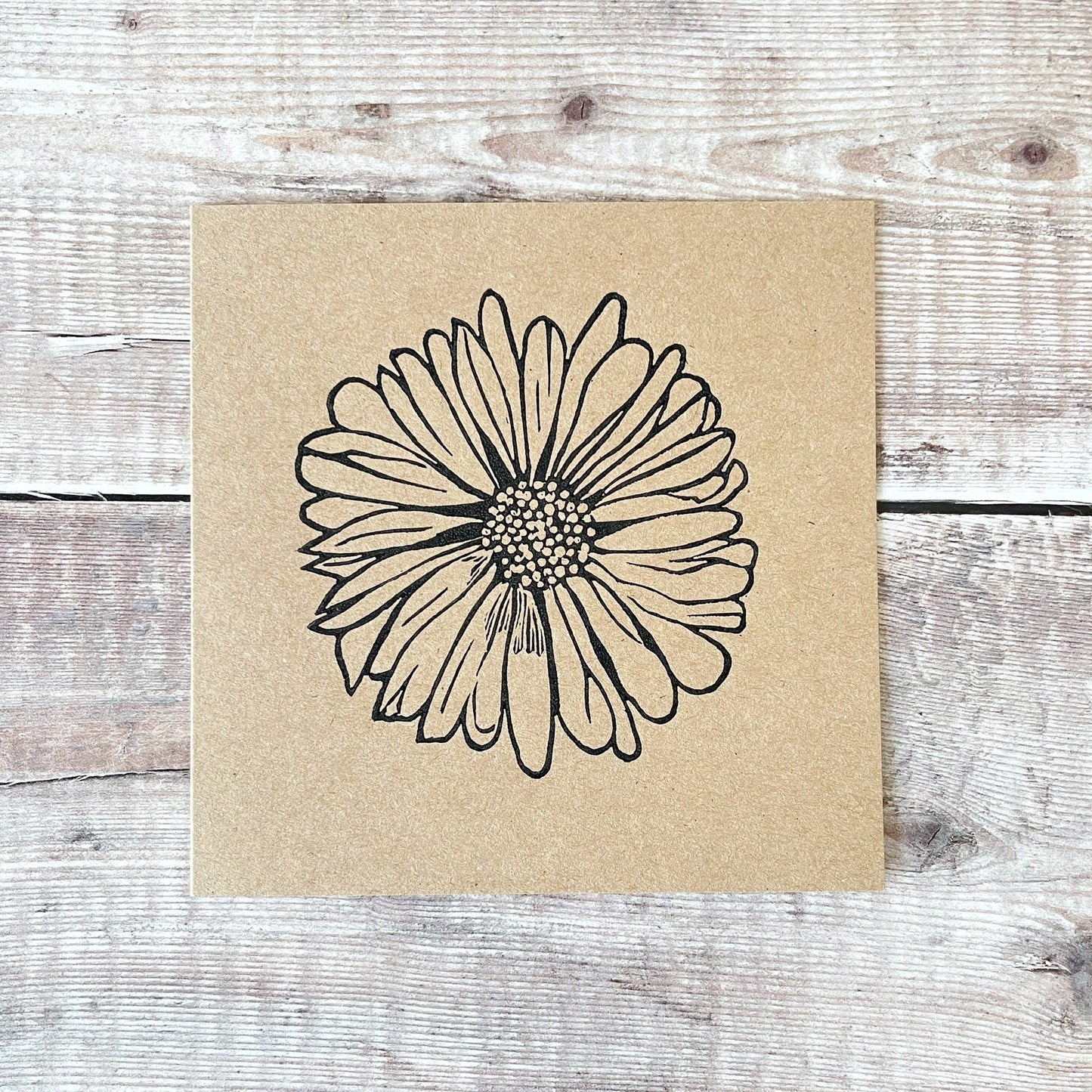 Kraft card with a black lino printed Daisy design laid on a wooden surface