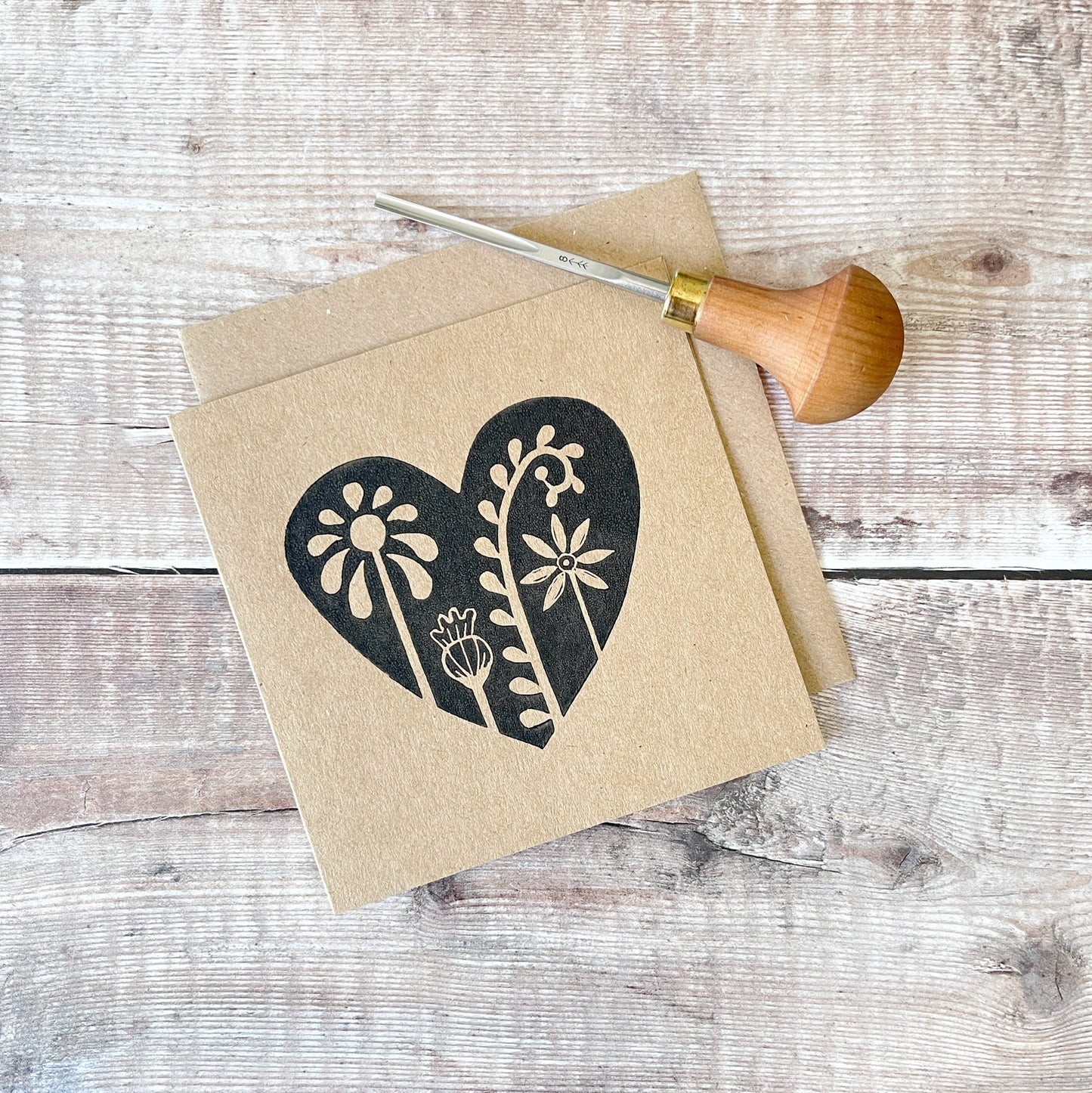 Floral heart design lino printed in black on a kraft card with a matching envelope. Laid on a wooden background the photo includes the lino cutting tool used.
