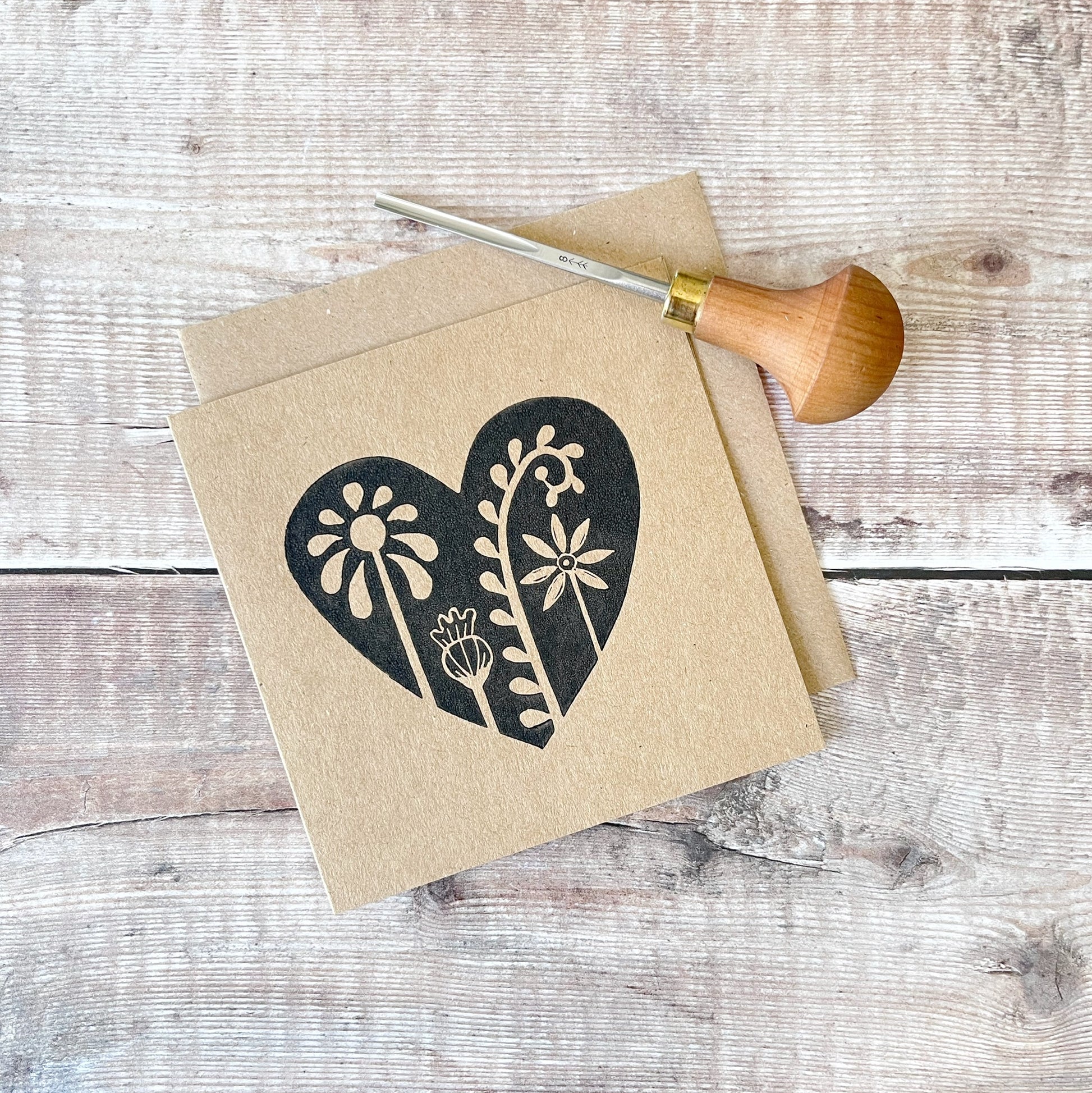 Floral heart design lino printed in black on a kraft card with a matching envelope. Laid on a wooden background the photo includes the lino cutting tool used.