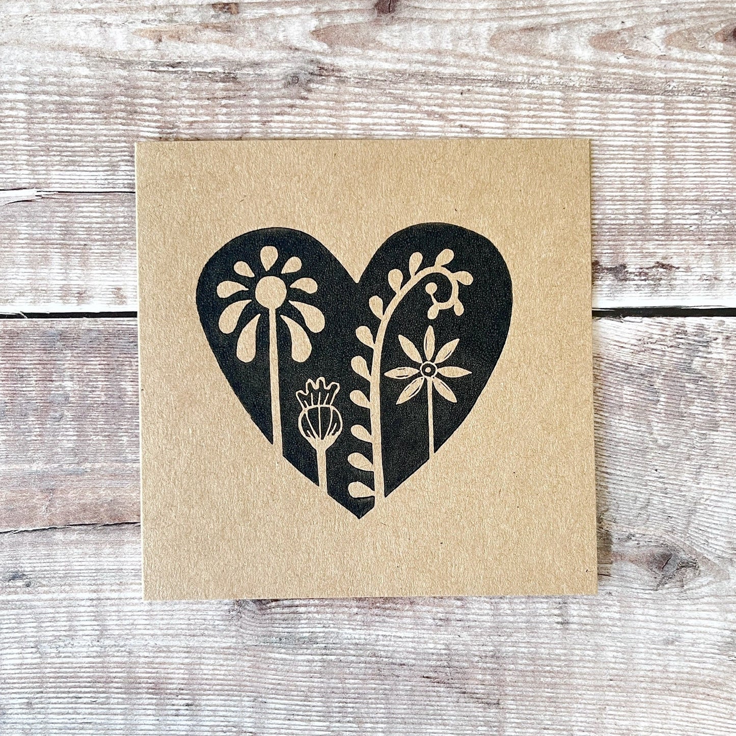 Floral heart design lino printed in black on kraft card and laid on a wooden background.