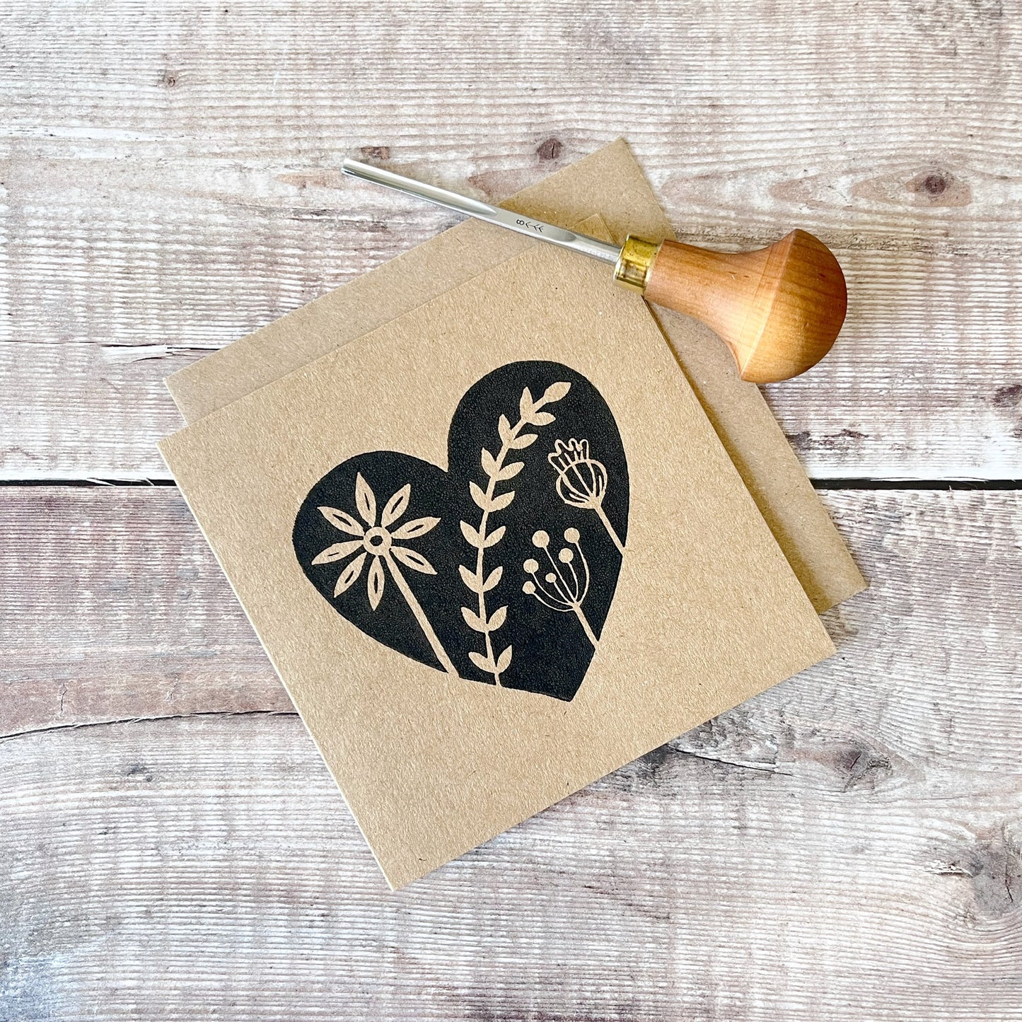 Black lino printed heart with floral design on kraft card with a matching envelope laid on a wooden surface. Photo includes the lino cutting tool used.