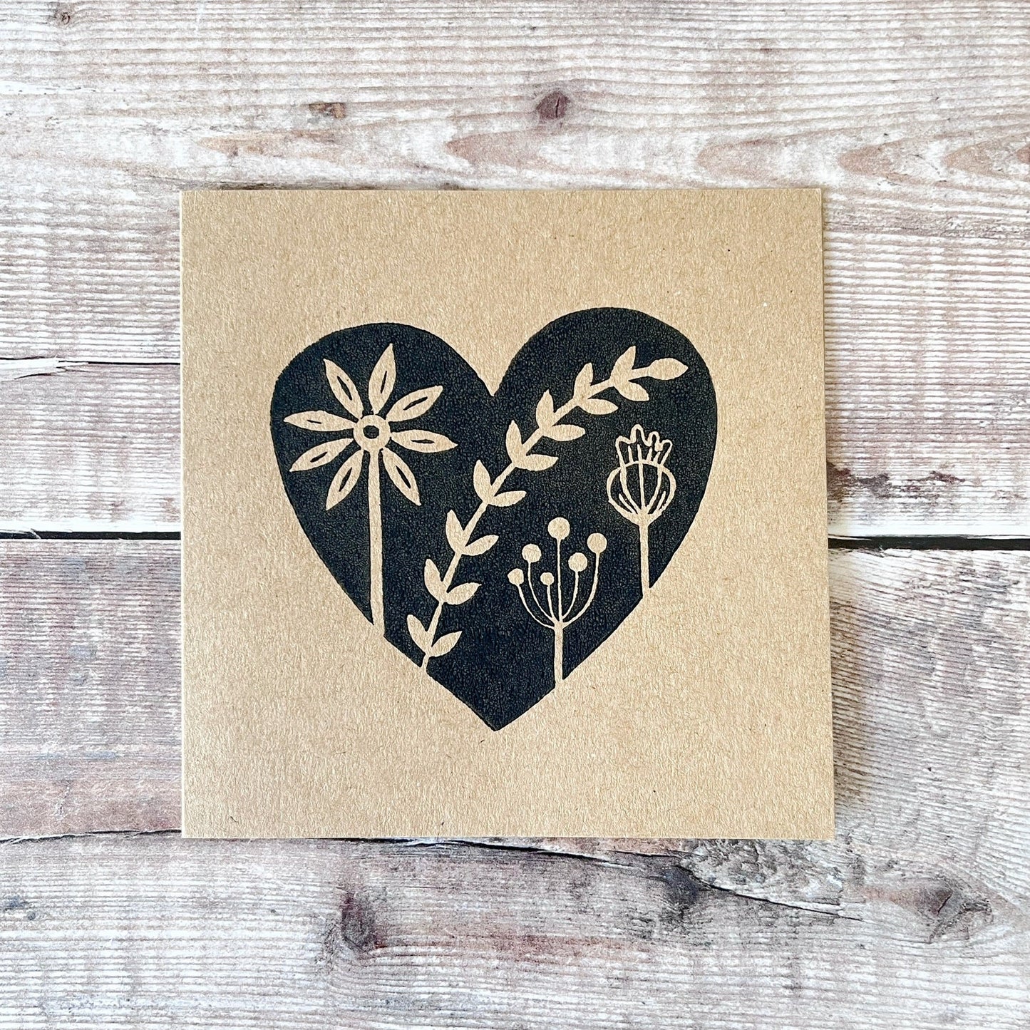 Kraft card with a black lino printed heart design featuring floral elements laid on a wooden surface