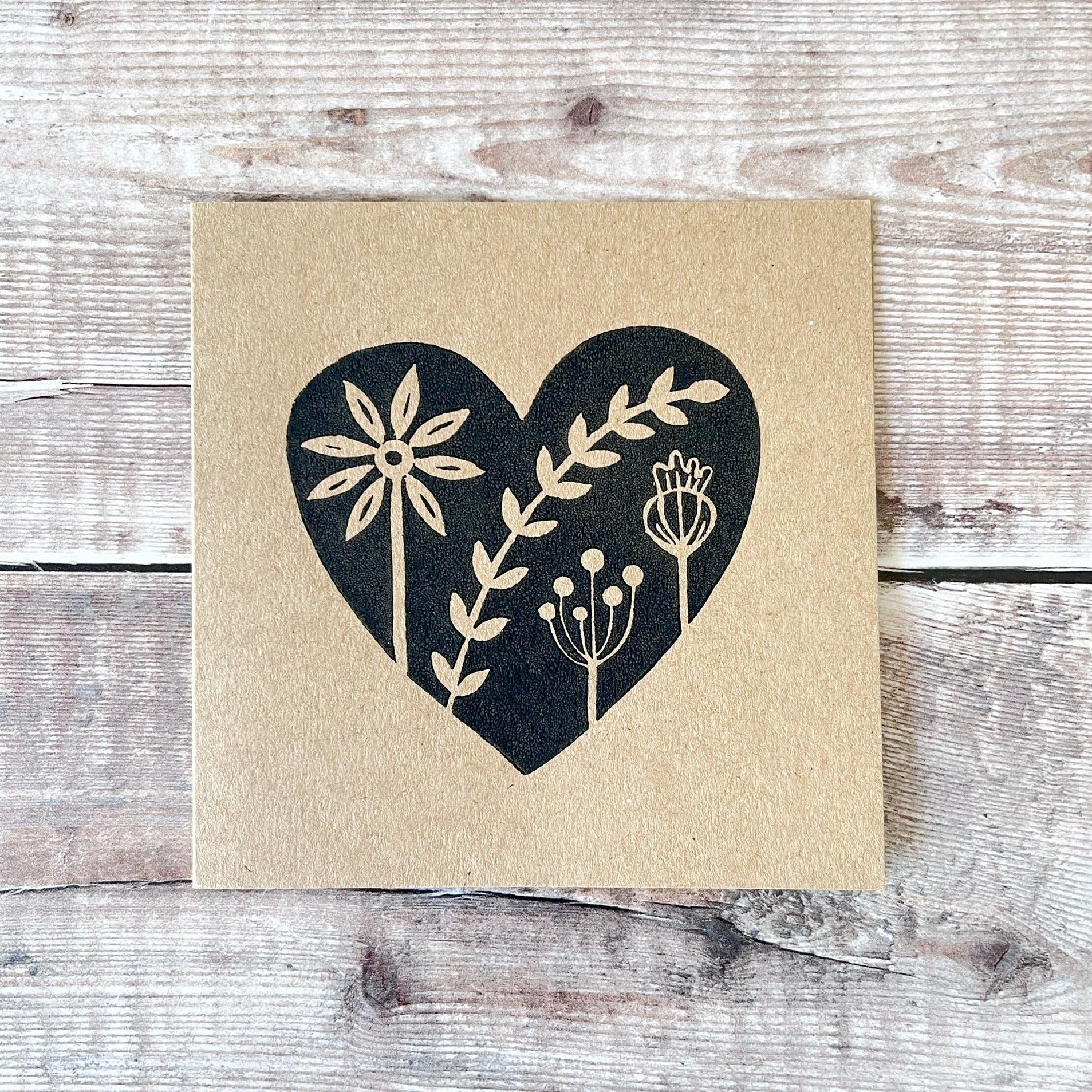 Kraft card with a black lino printed heart design featuring floral elements laid on a wooden surface