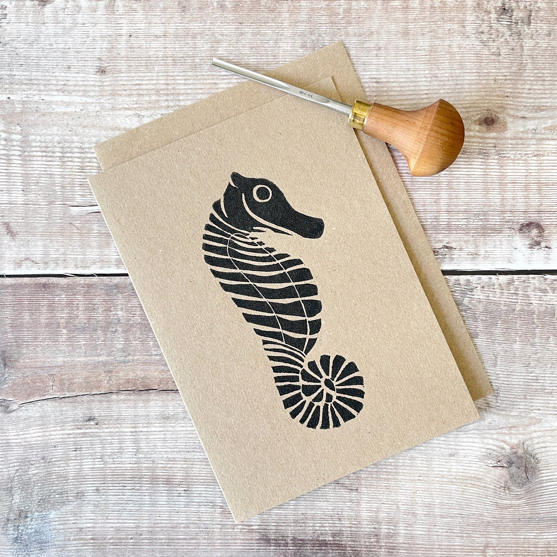 Black lino printed seahorse on a kraft card with a matching envelope laid on a wooden background. Photo also includes lino cutting tool that was used.