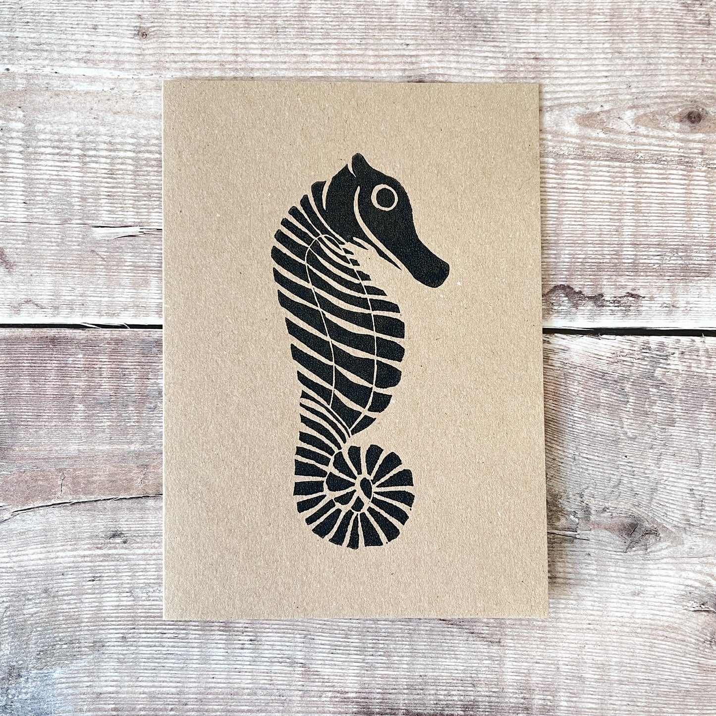 Black lino printed seahorse on a kraft card laid on a wooden background.