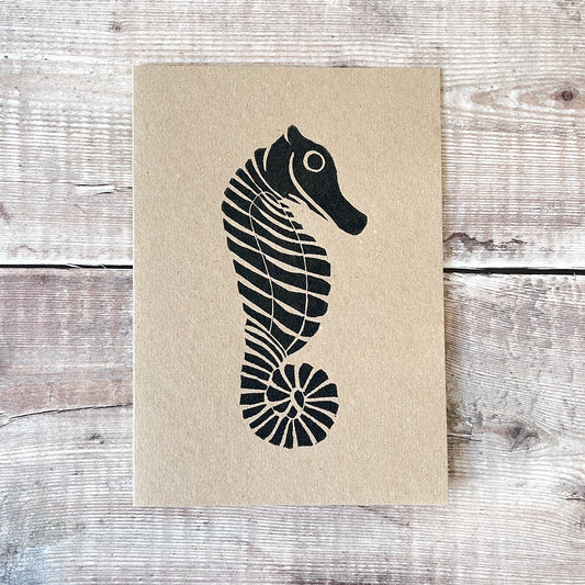 Black lino printed seahorse on a kraft card laid on a wooden background.