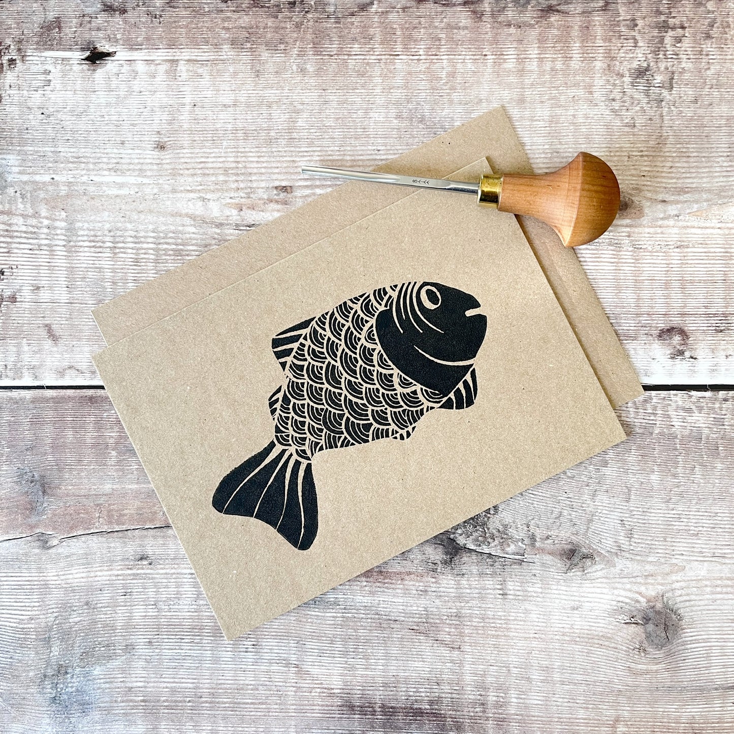 Black lino print of a fish illustration on kraft card with a matching envelope laid on a wooden surface. Photo includes the lino cutting tool used.