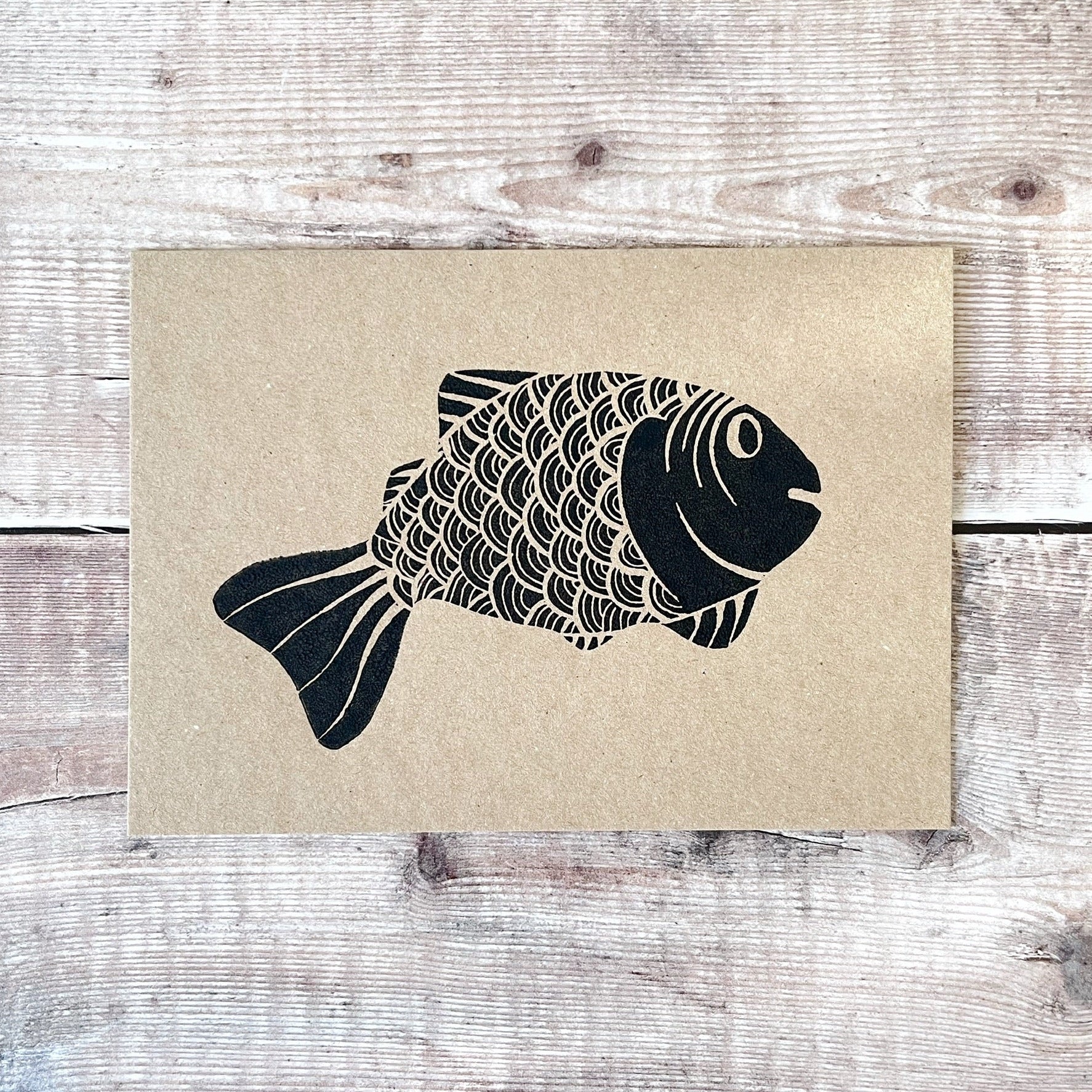 Black lino print of a fish illustration on a kraft card laid wooden surface